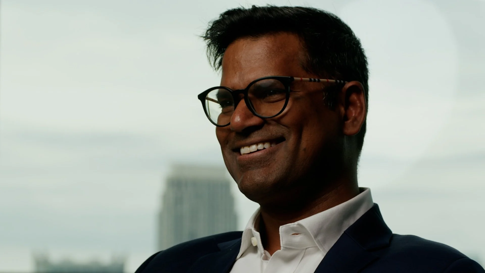 A man smiling, wearing glasses and a formal suit, standing outdoors with a cityscape and cloudy sky in the background.