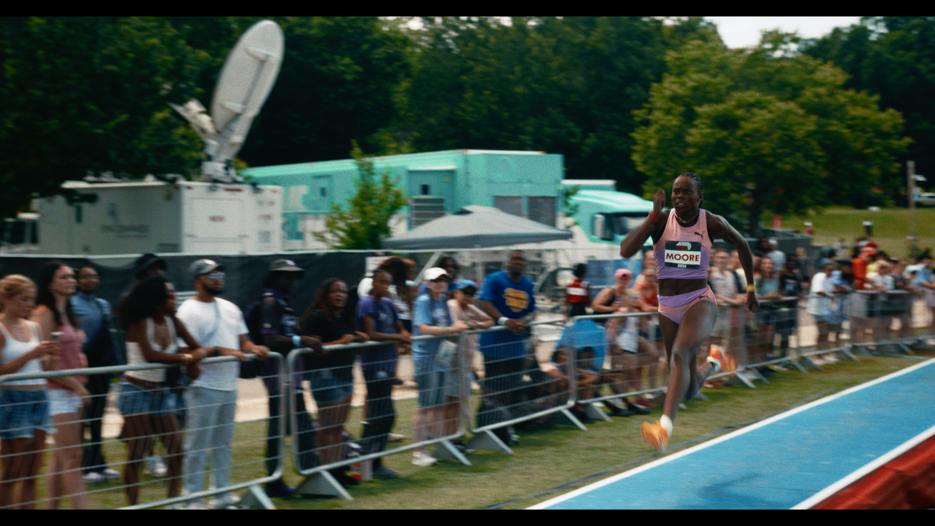 A female sprinter running on a track during a race, with spectators watching from behind a barrier and large vehicles and trees in the background.