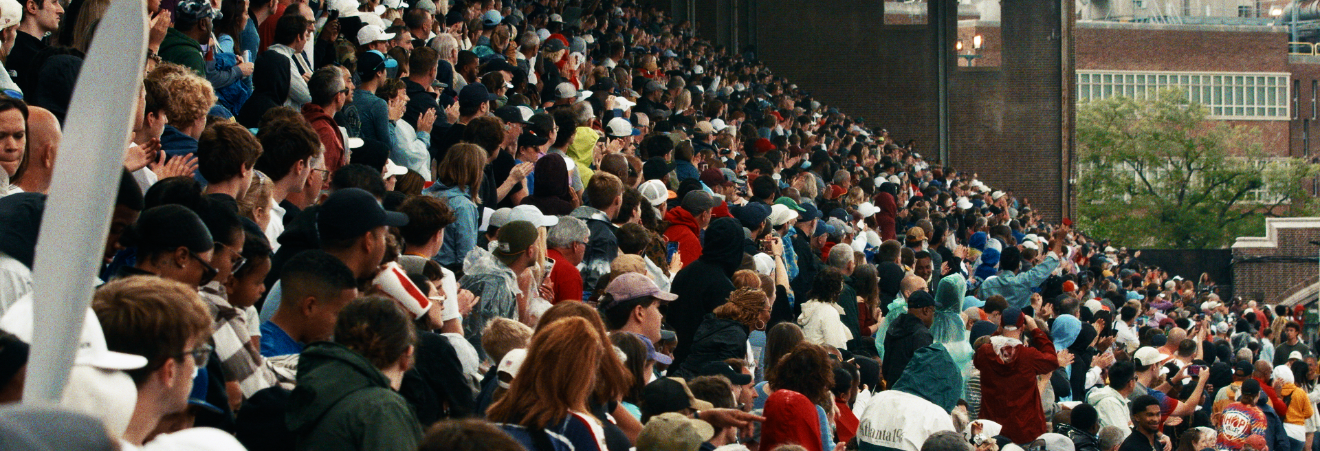A large crowd of people gathered in a stadium bleacher, some clapping and cheering, with diverse ages and backgrounds, wearing casual clothing and hats.