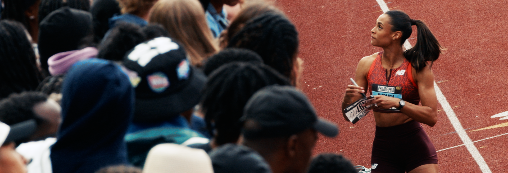Female athlete in red sports bra and black shorts on running track, holding a race bib and a pen, with an audience watching.
