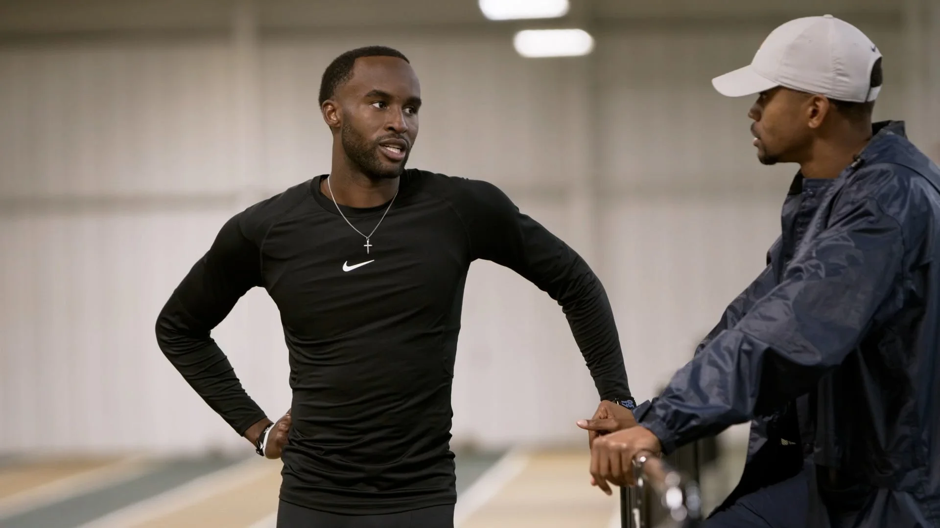 Two men having a conversation in an indoor sports facility, one in a black Nike shirt with a cross necklace, and the other in a gray cap and navy jacket.