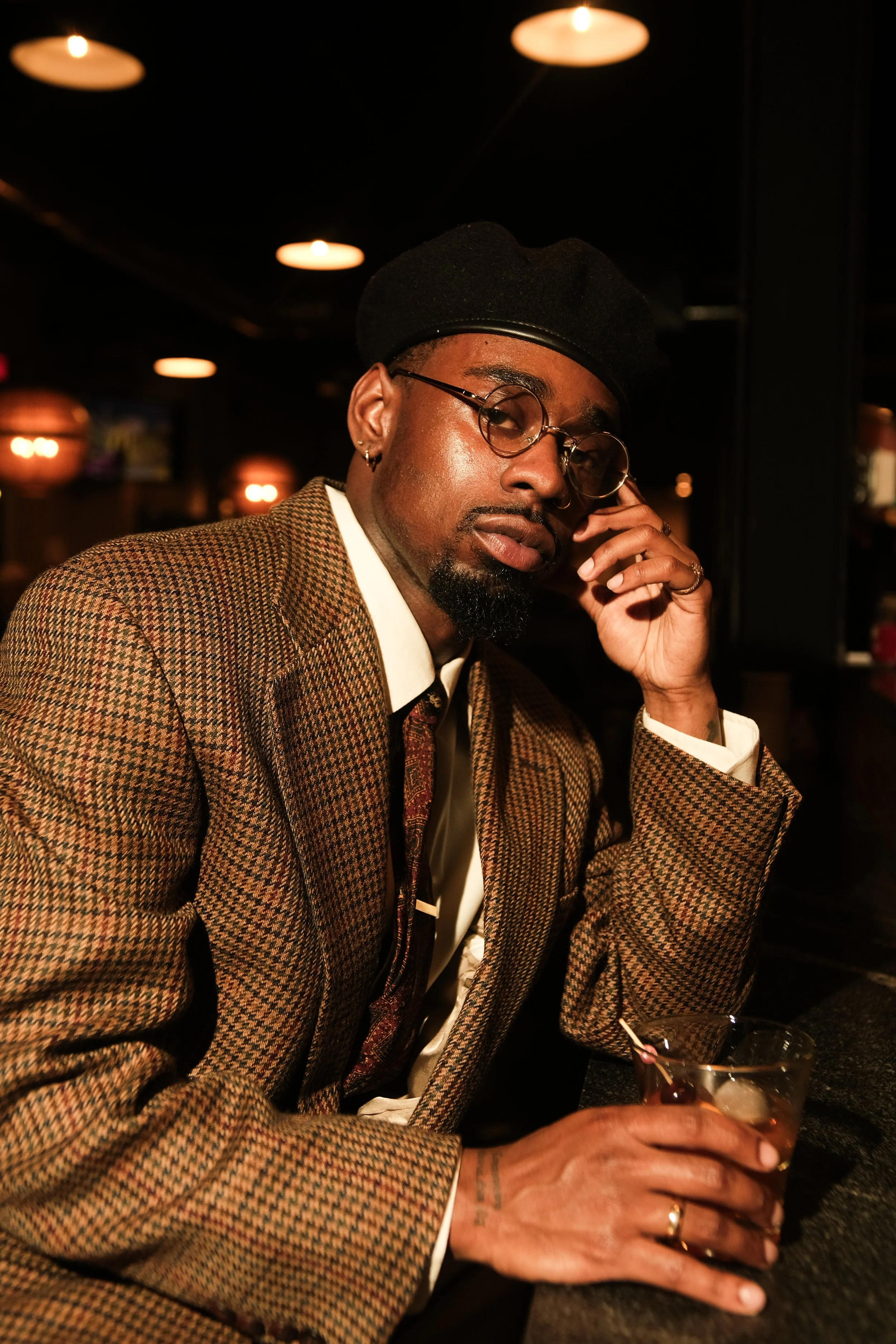 A man in a brown checkered suit, white shirt, and patterned tie sitting in a dimly lit bar or lounge, holding a drink with ice and a straw, wearing glasses and a black beret, with a serious expression.