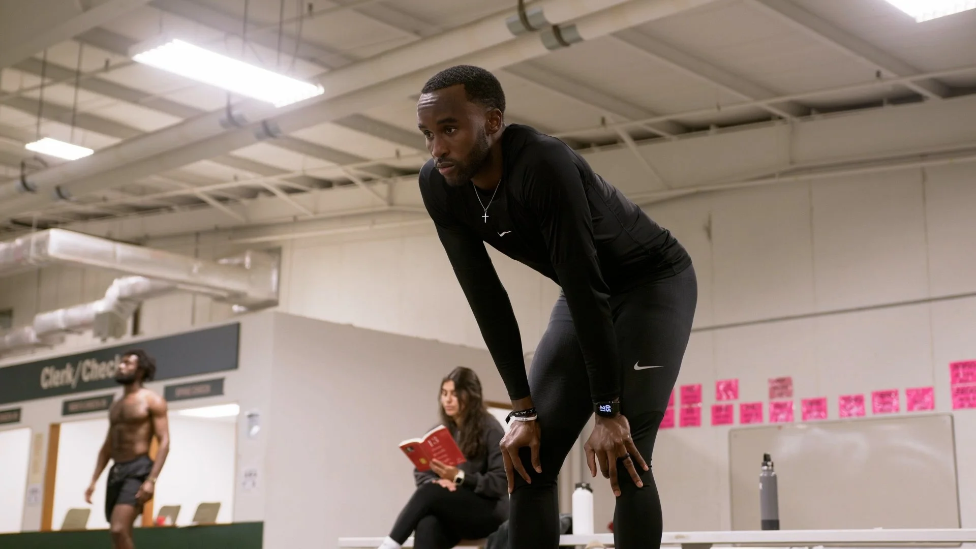 A man in black athletic clothing bent over resting with his hands on his knees in a gym, with a woman seated behind him reading a book and a shirtless man in the background.