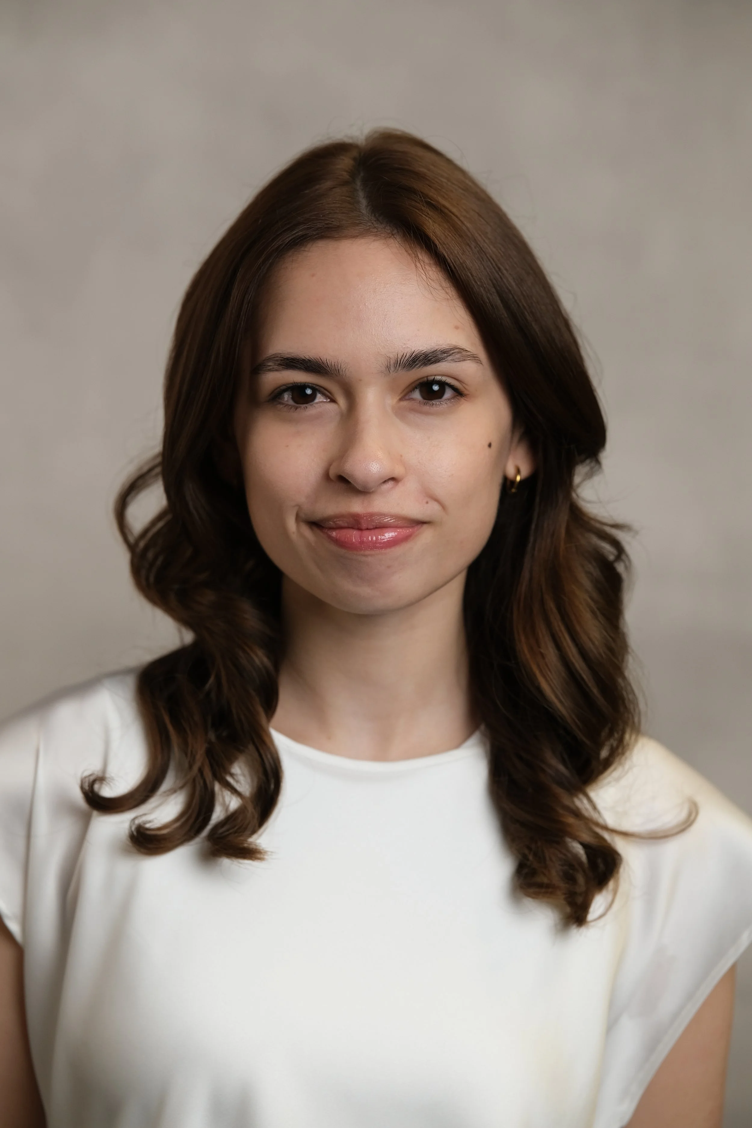 A woman with brown, wavy hair and light skin wearing a white top and gold earrings, smiling slightly against a neutral background.