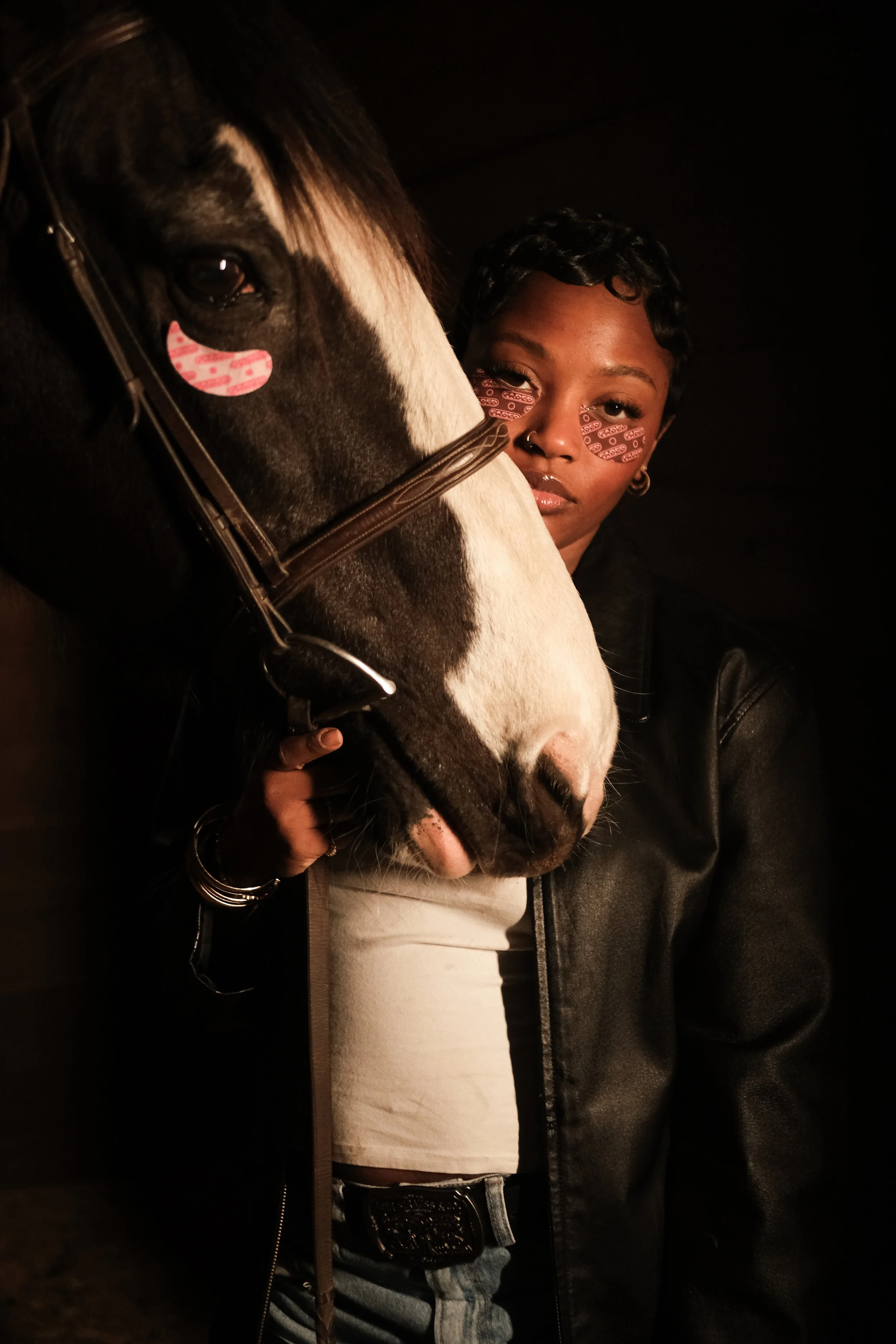 A person with curled short hair, wearing a black leather jacket and white top, standing next to a horse with black and white markings, in a dark setting.