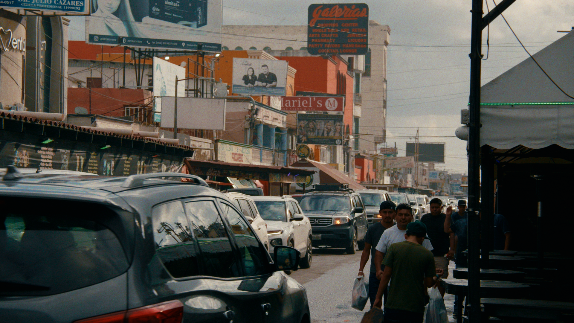 Street scene with parked cars, pedestrians walking, and colorful buildings with signs and billboards in the background.
