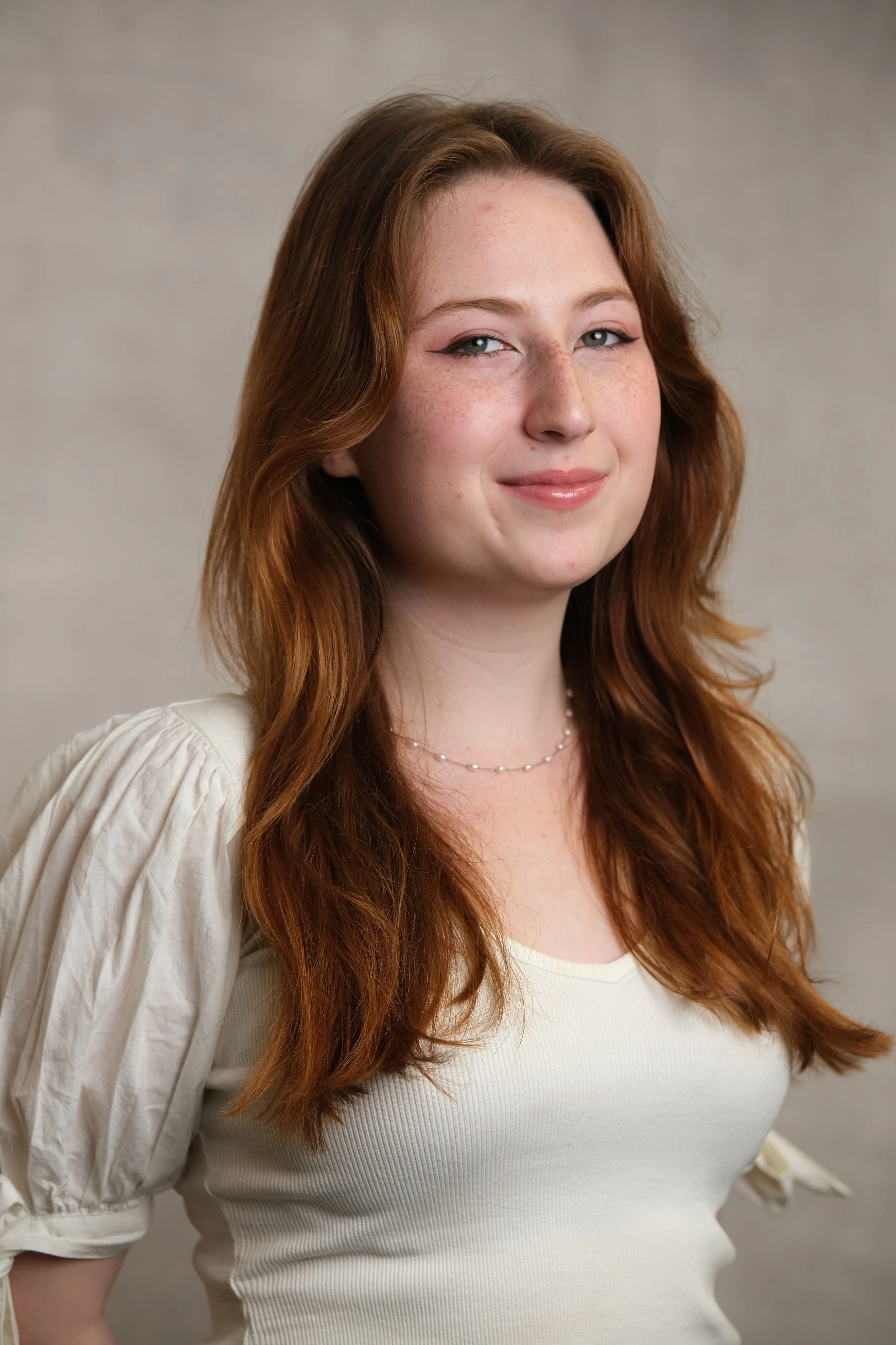 A young woman with long, wavy red hair and blue eyes, wearing a cream-colored top with puffed sleeves and a delicate necklace, smiling softly at the camera against a plain background.