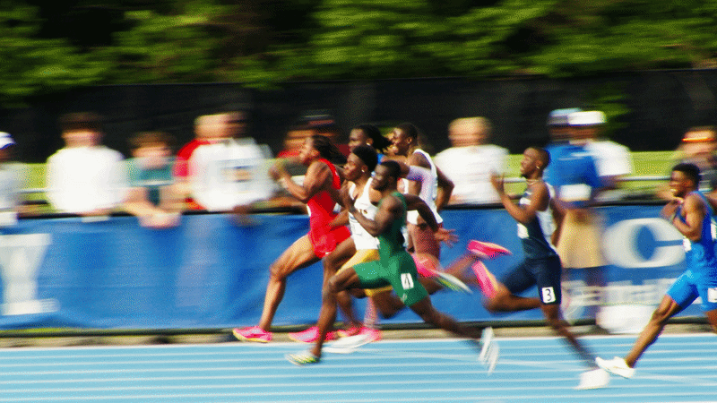 Group of runners competing in a track race, with spectators blurred in the background.