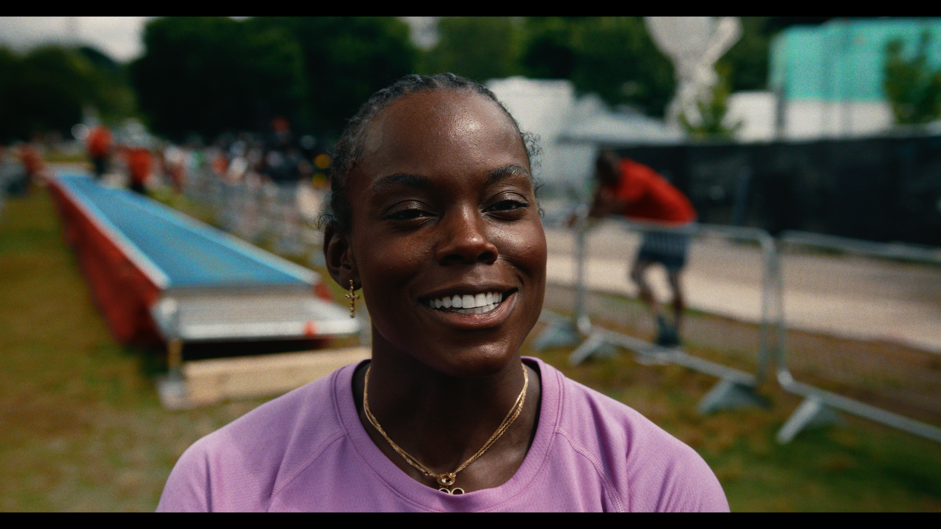 Smiling woman with long braided hair, wearing a purple athletic shirt and jewelry, outdoors at a track and field event with blurred background of people, track, and trees.