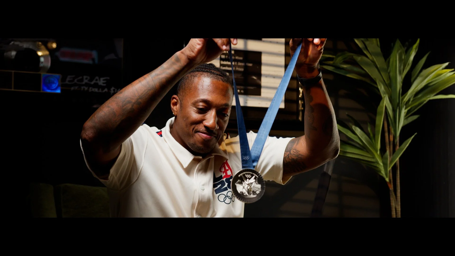 Athlete proudly displays her silver medal from the Olympic Games, wearing a white shirt with a logo, with a green plant and dark background behind her.