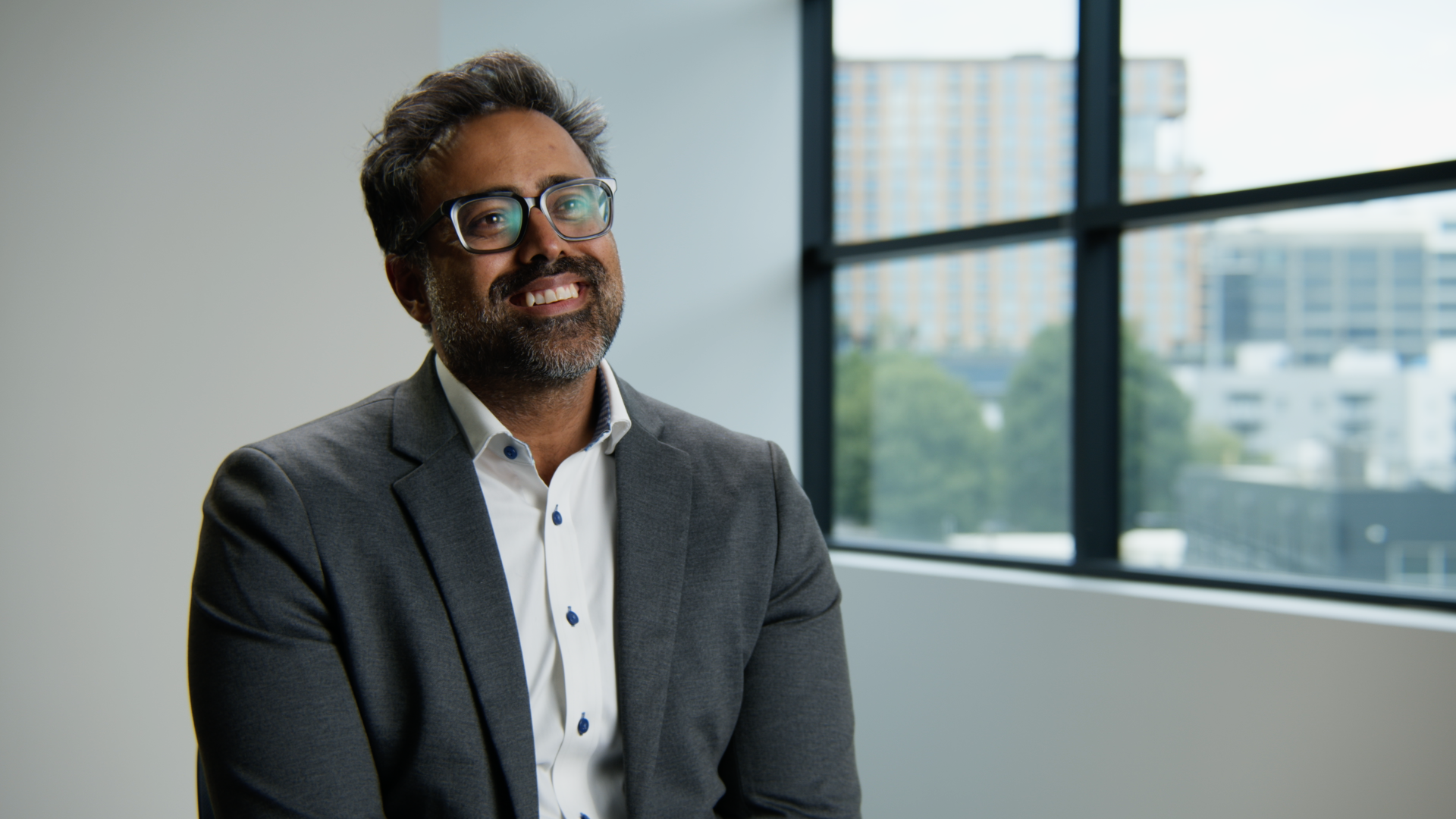 A middle-aged man with glasses, a beard, and dark hair, wearing a gray blazer and white shirt, smiling while sitting in a modern office near large windows with a cityscape background.