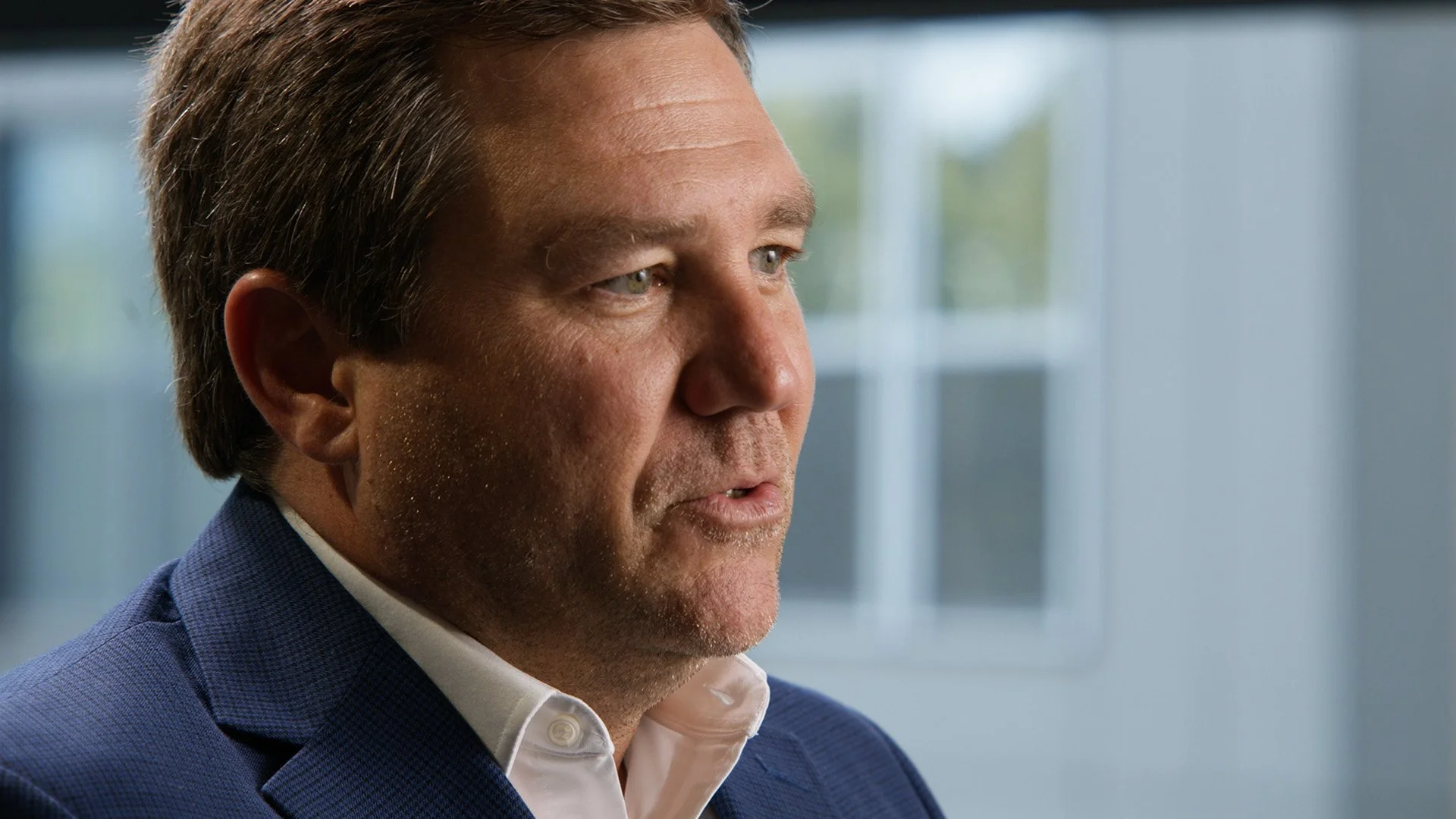 Close-up of a middle-aged man with brown hair, wearing a blue suit and white shirt, sitting indoors near a window.