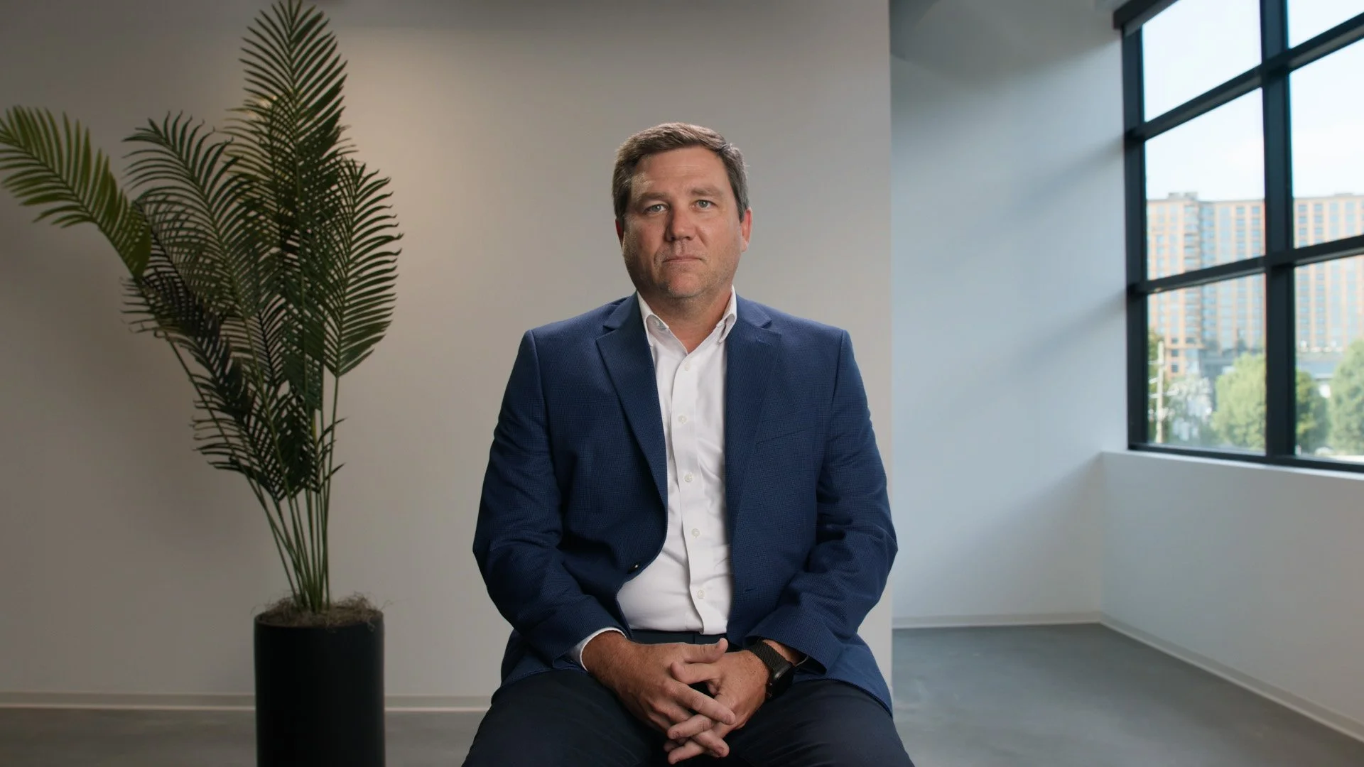A man in a blue suit and white shirt sitting in a bright office with a large window and a potted plant nearby.