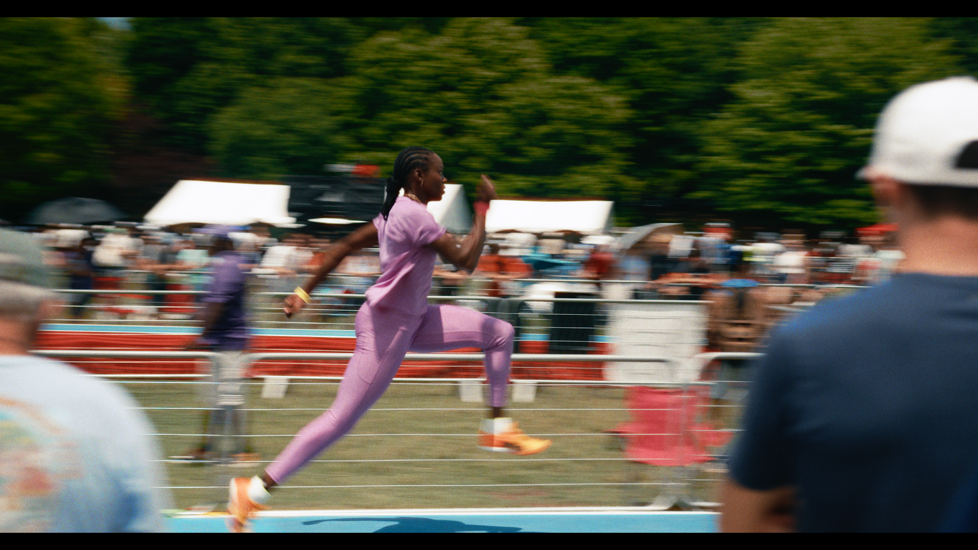 A female runner in a purple outfit sprinting on a track during a race, with spectators and trees in the background, and blurred figures in the foreground.