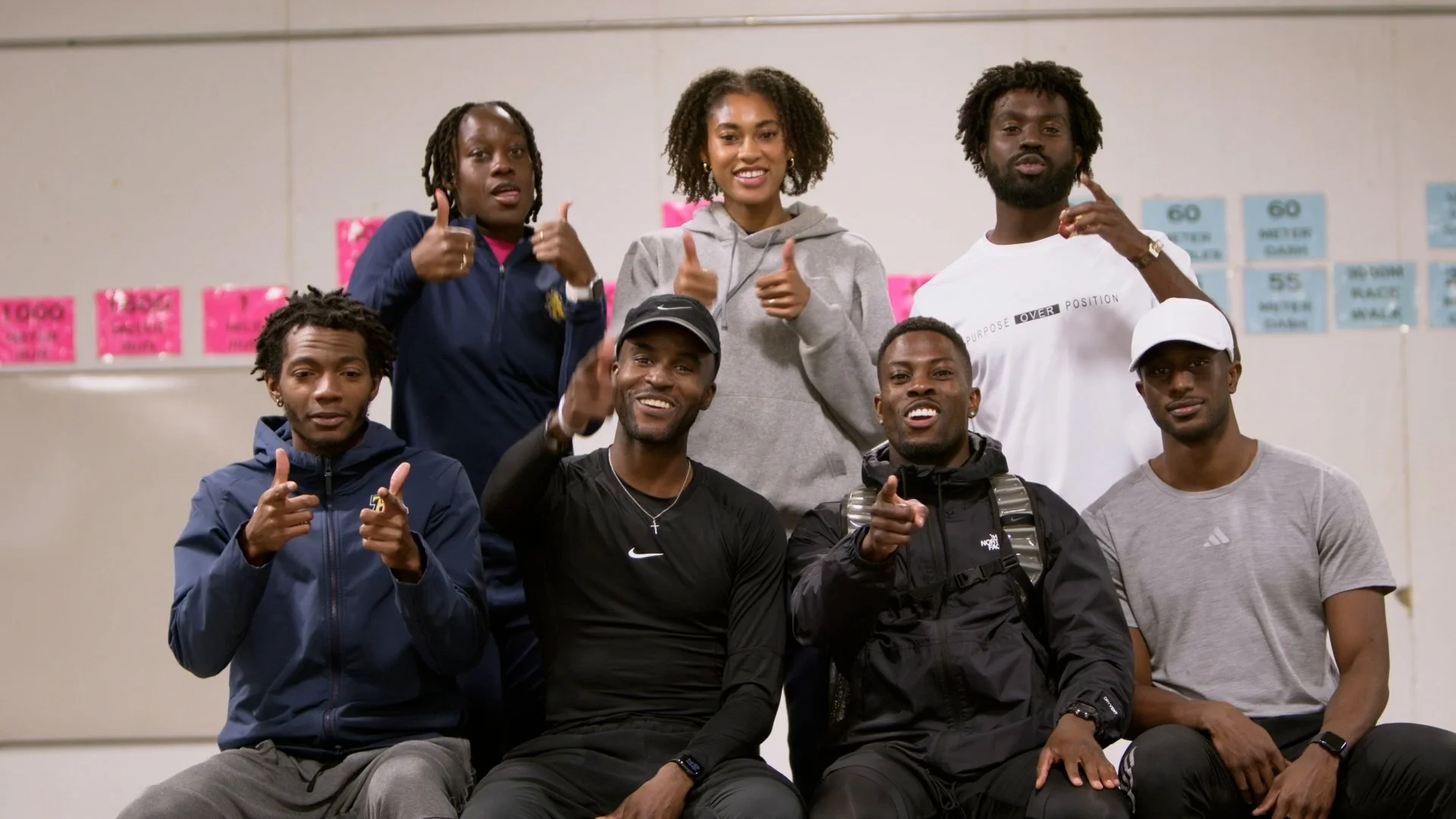 Group of seven young adults in casual athletic attire posing together indoors, some showing thumbs-up and pointing at the camera, with pink and blue posters on the wall in the background.