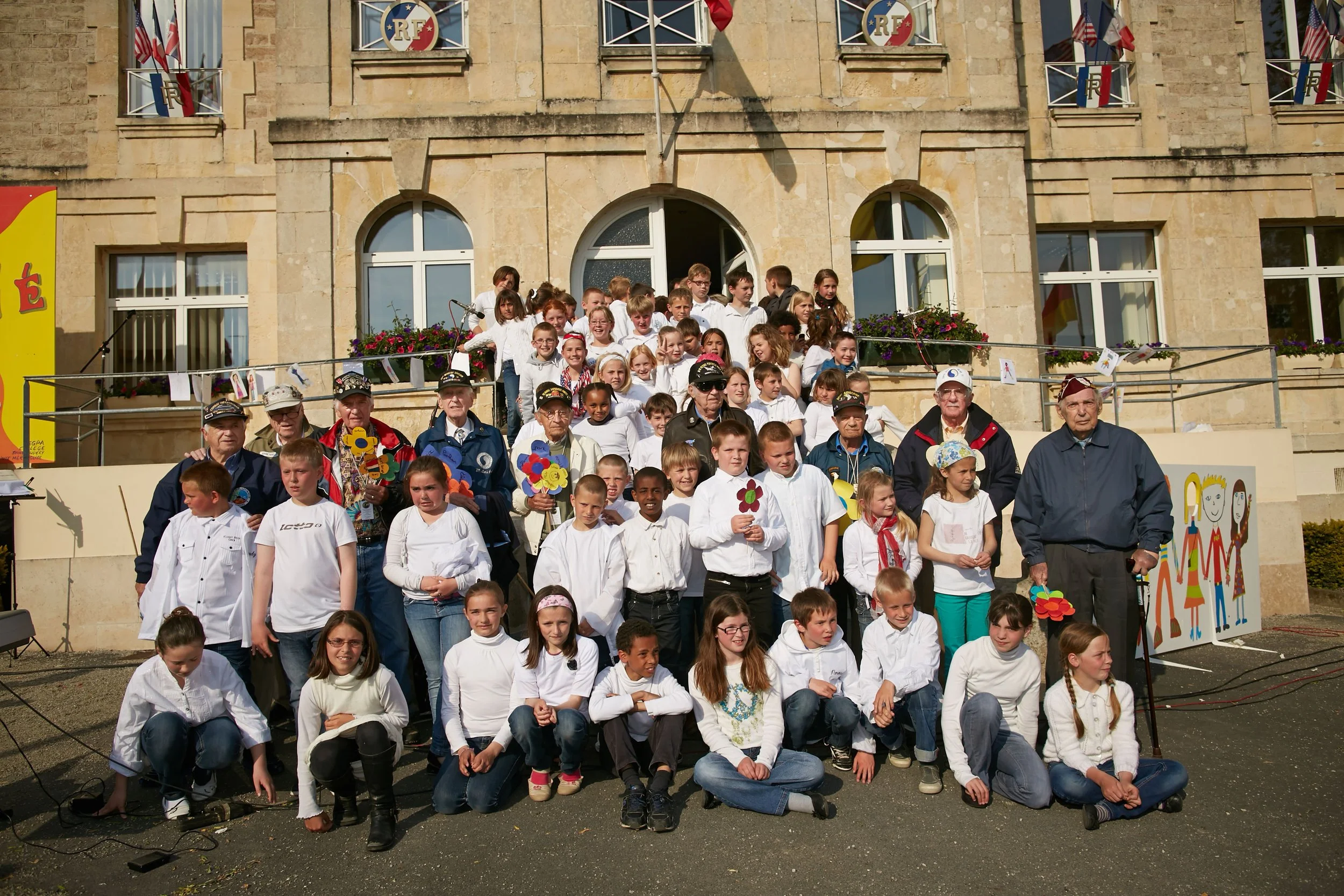 Group of children and elderly people gathered on a stage in front of a building decorated with flags and banners for a school or community event.