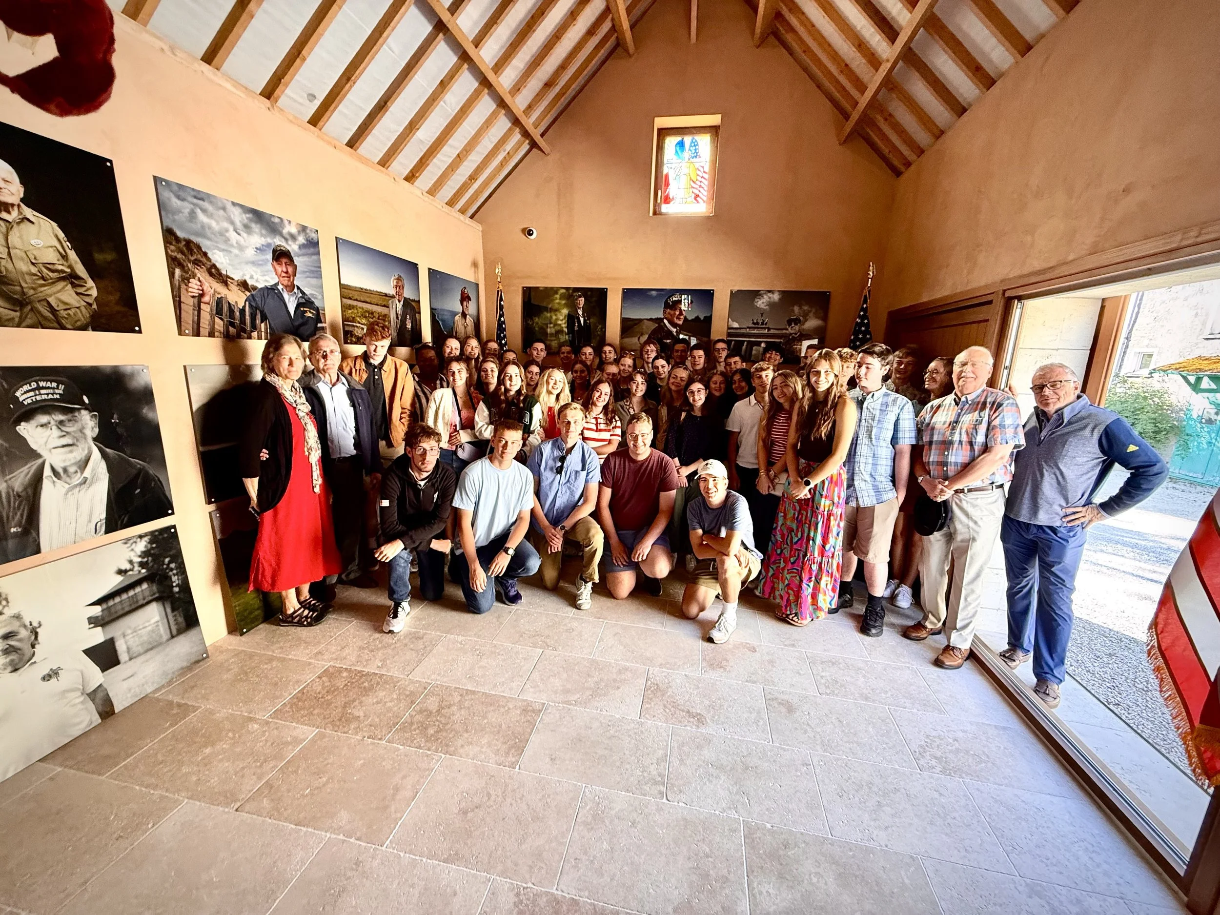 Group of people gathered inside a room with paintings of veterans on the wall, large window to the right, and a stained glass window on the back wall.
