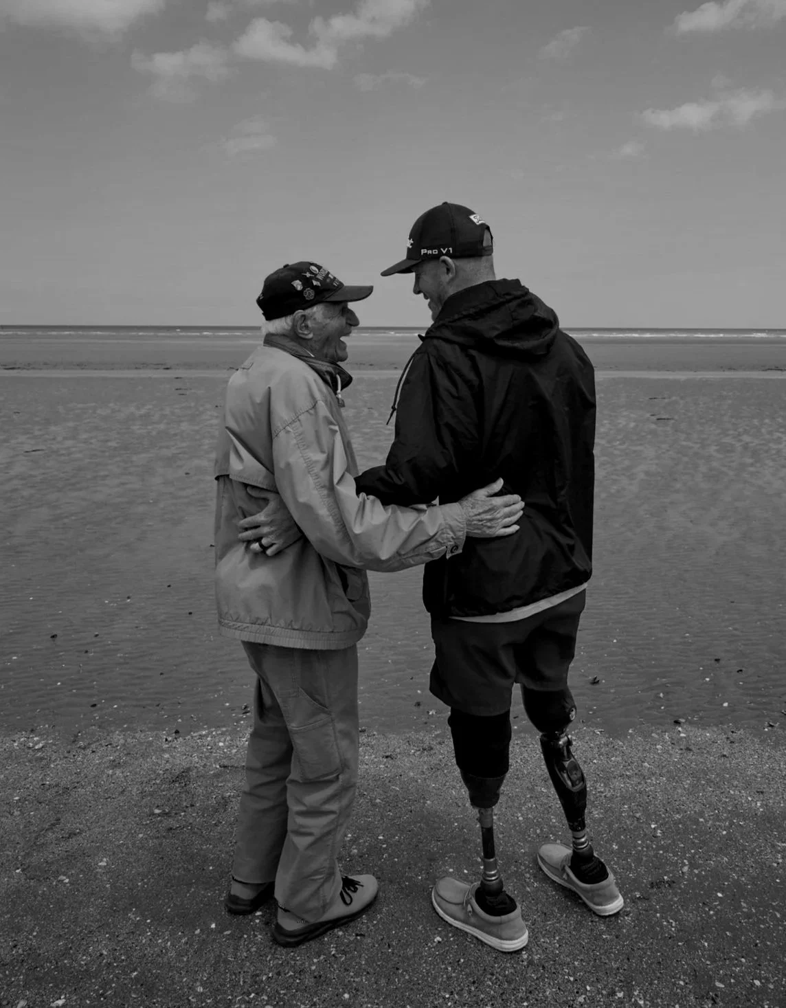 An elderly man and a younger man with a prosthetic leg are holding hands and smiling at each other on a beach, with the ocean in the background.