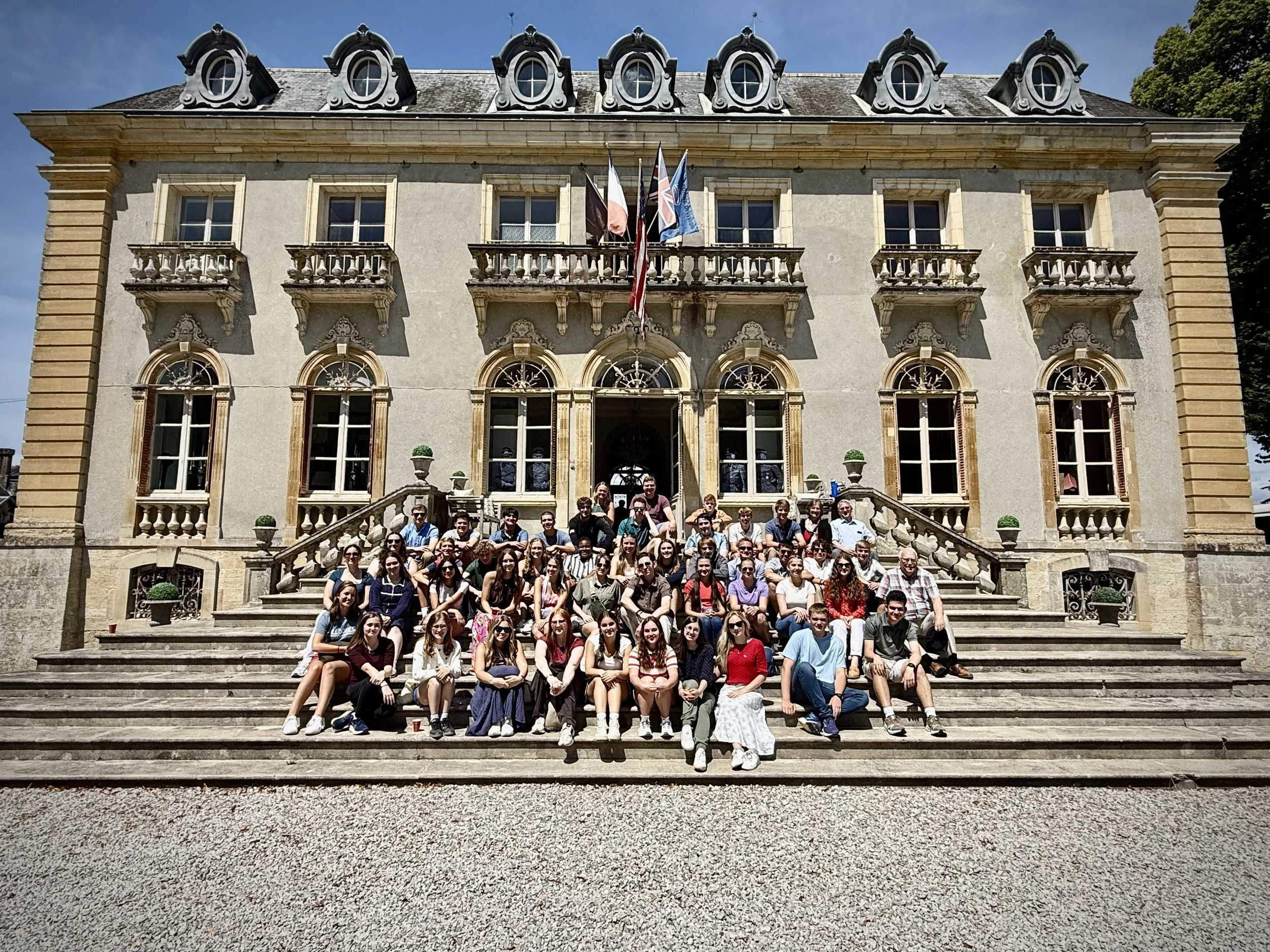 Large group of people sitting on the steps of a historic building with European architecture, arched windows, and flags flying above the entrance.