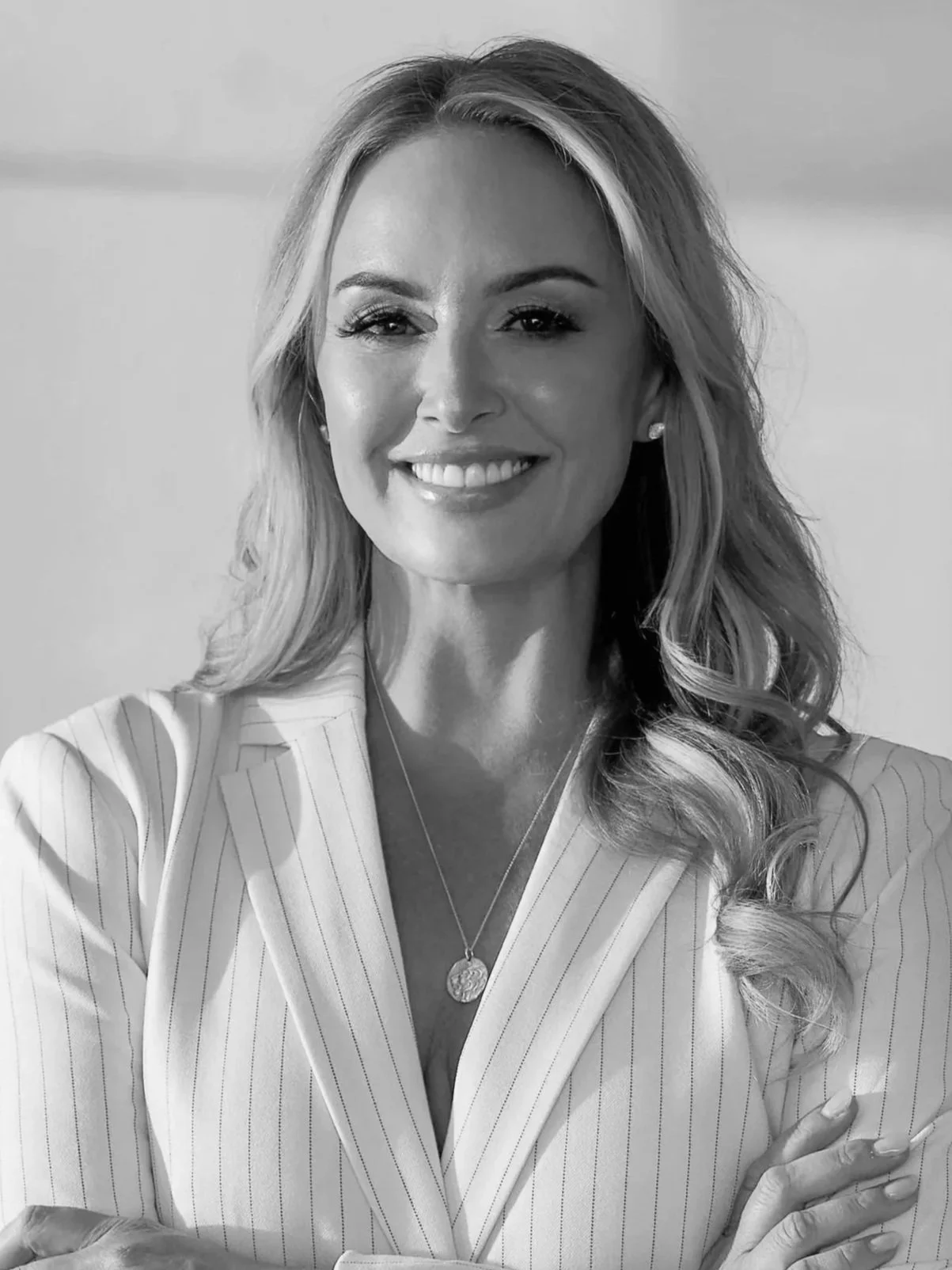 A black and white photo of a woman with long dark hair, smiling faintly, wearing a white top and a necklace with a pearl pendant.