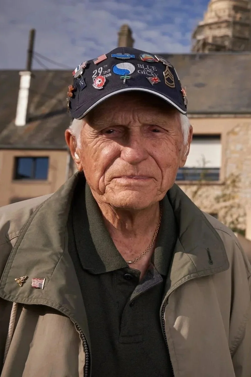 An elderly man wearing a black cap decorated with military pins and patches, a green jacket, and a black polo shirt, standing outside in front of a building with a chimney and a cloudy sky.