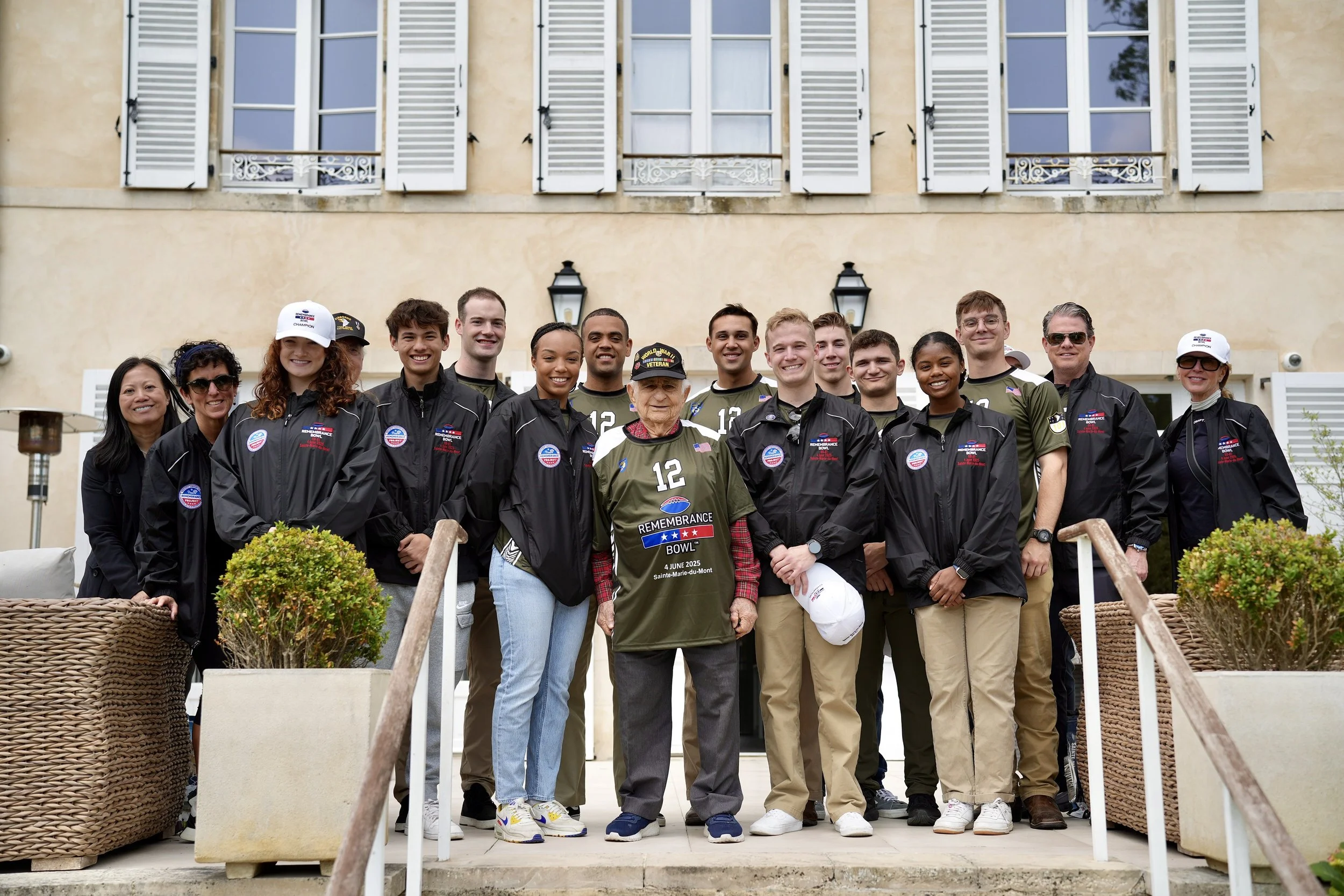 Group photo of young people and two older individuals wearing commemorative jackets and shirts, standing outside a building with beige walls and white window shutters, celebrating a remembrance event.