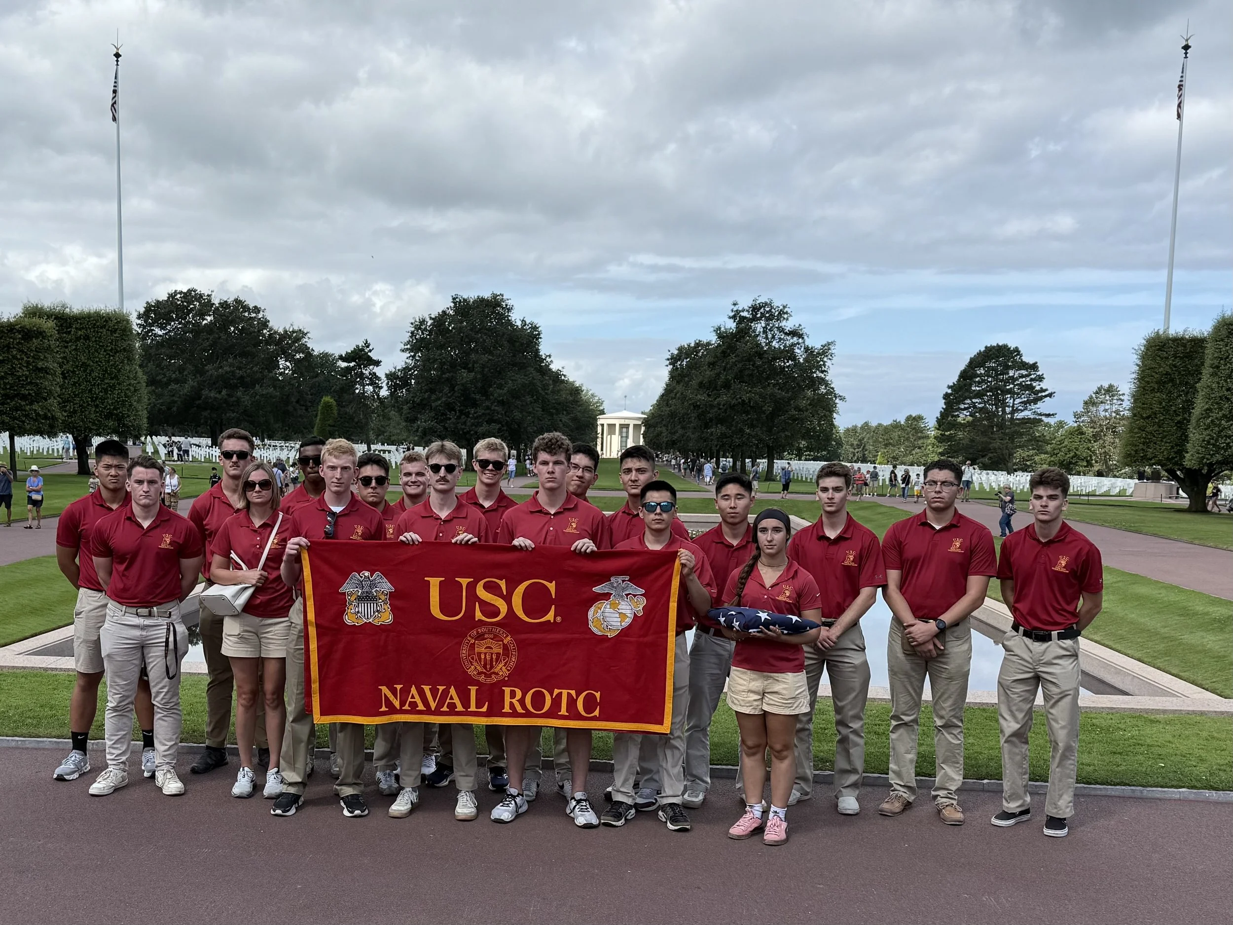 Group of young people in red shirts standing outdoors, holding a red banner that reads 'USC NAVAL ROTC', with a flag in the center, in a park-like setting with trees and flags in the background.