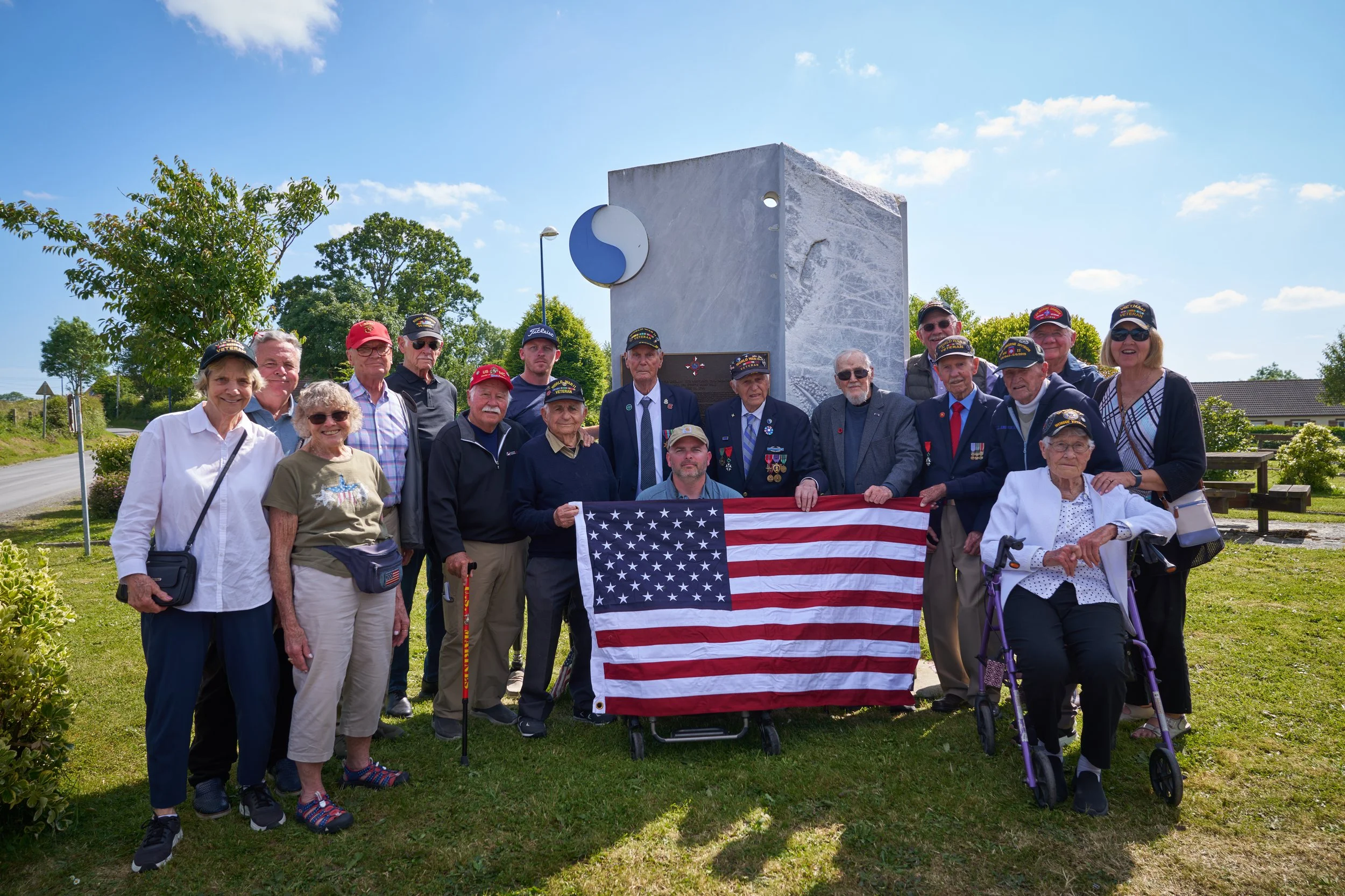 Group of elderly veterans and supporters gathered outdoors around a memorial, some wearing medals and hats, holding an American flag.