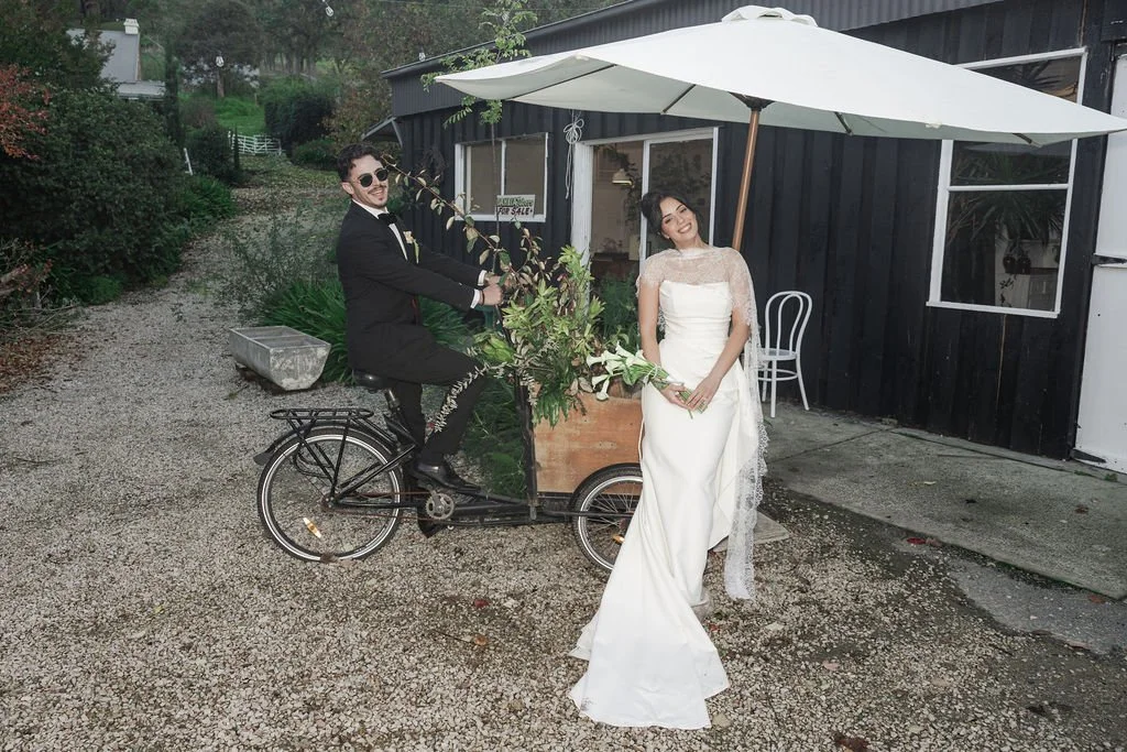 Bride and Groom having fun on a bike outside a black building in a jane hill gown with a lace vintage scarf