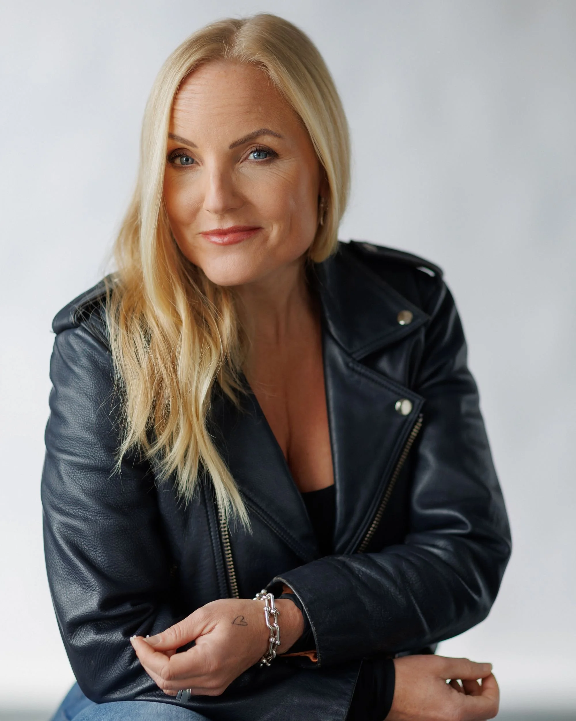 A woman with long blonde hair wearing a black leather jacket, smiling, with crossed arms, light background.