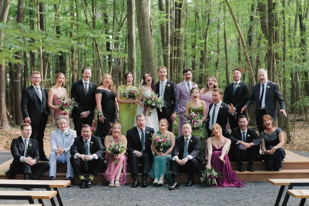 Group of people in formal attire, resembling wedding party, smiling and laughing on a wooden platform in a wooded outdoor setting.