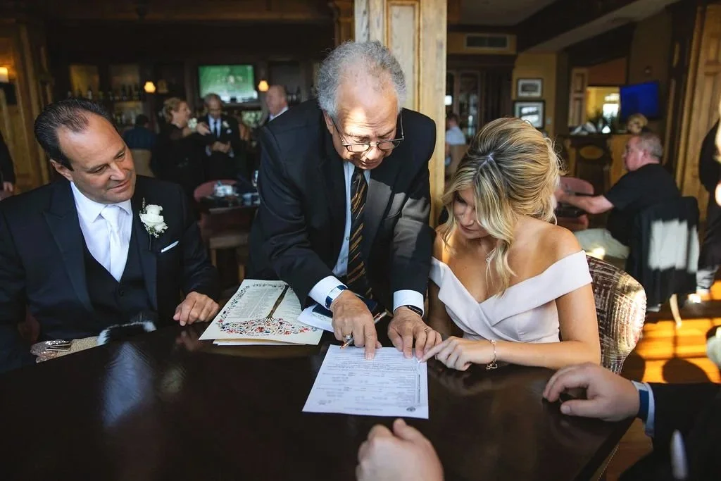 A bride and groom sitting at a table during their wedding ceremony, with an officiant reviewing the marriage license or vows.