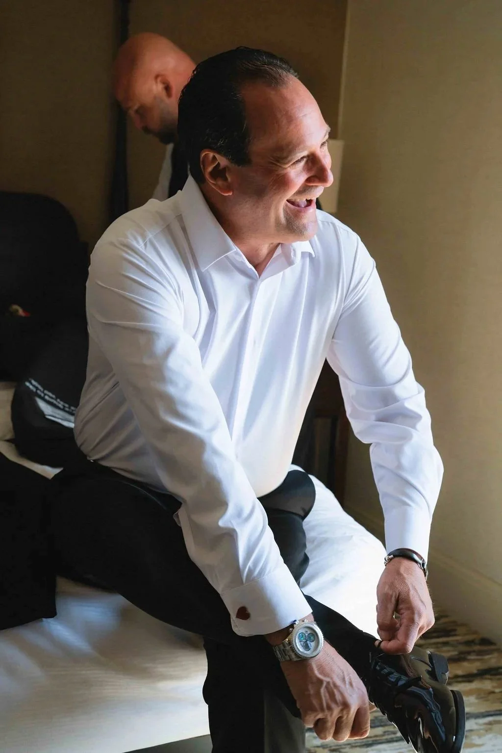A man in a white dress shirt sitting on a bed, smiling, as he ties his black dress shoes. Another man is in the background.