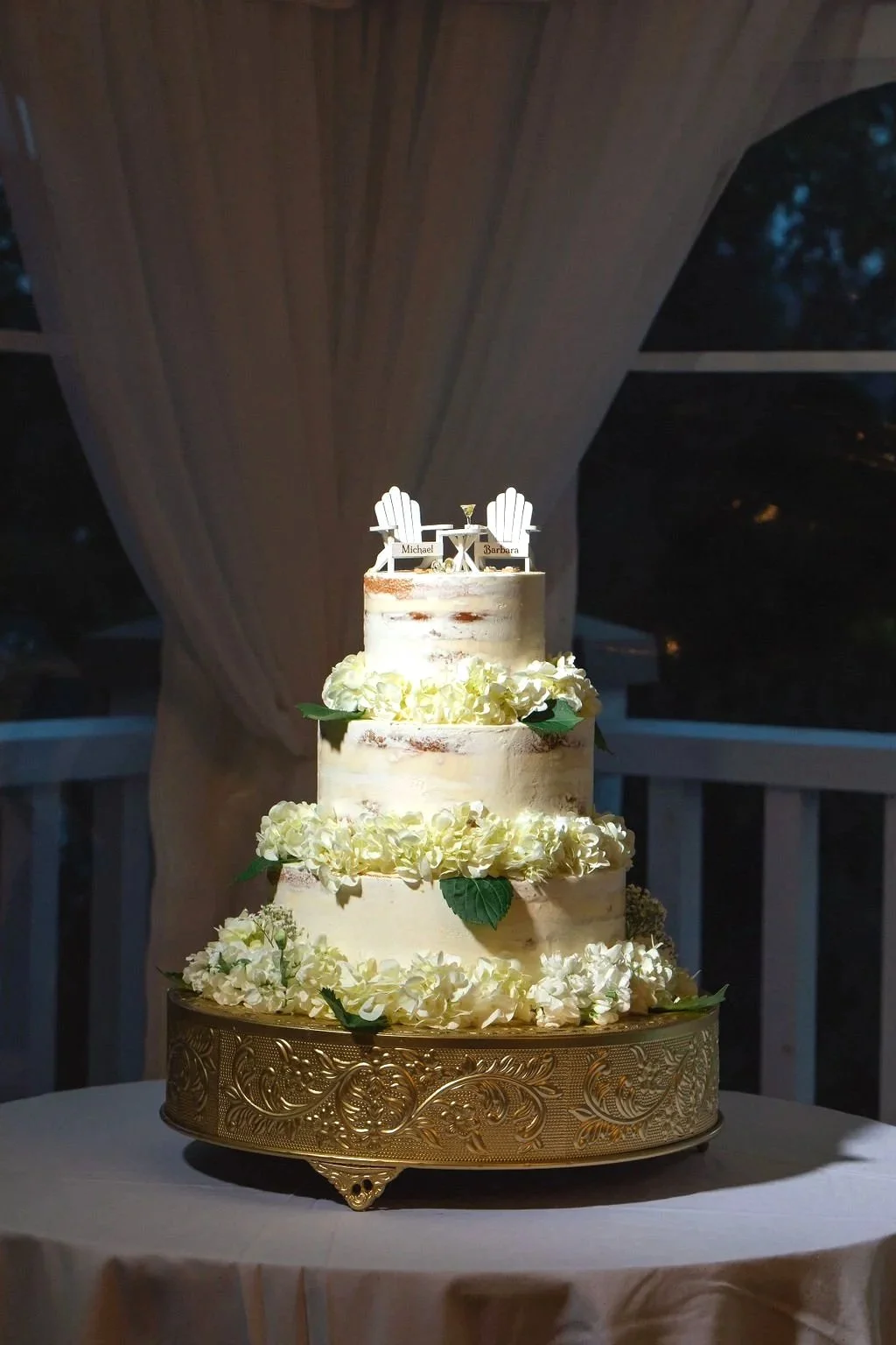 Four-tier wedding cake decorated with white flowers and green leaves, topped with miniature Adirondack chairs labeled 'Michael' and 'Barbara,' and a small drink.