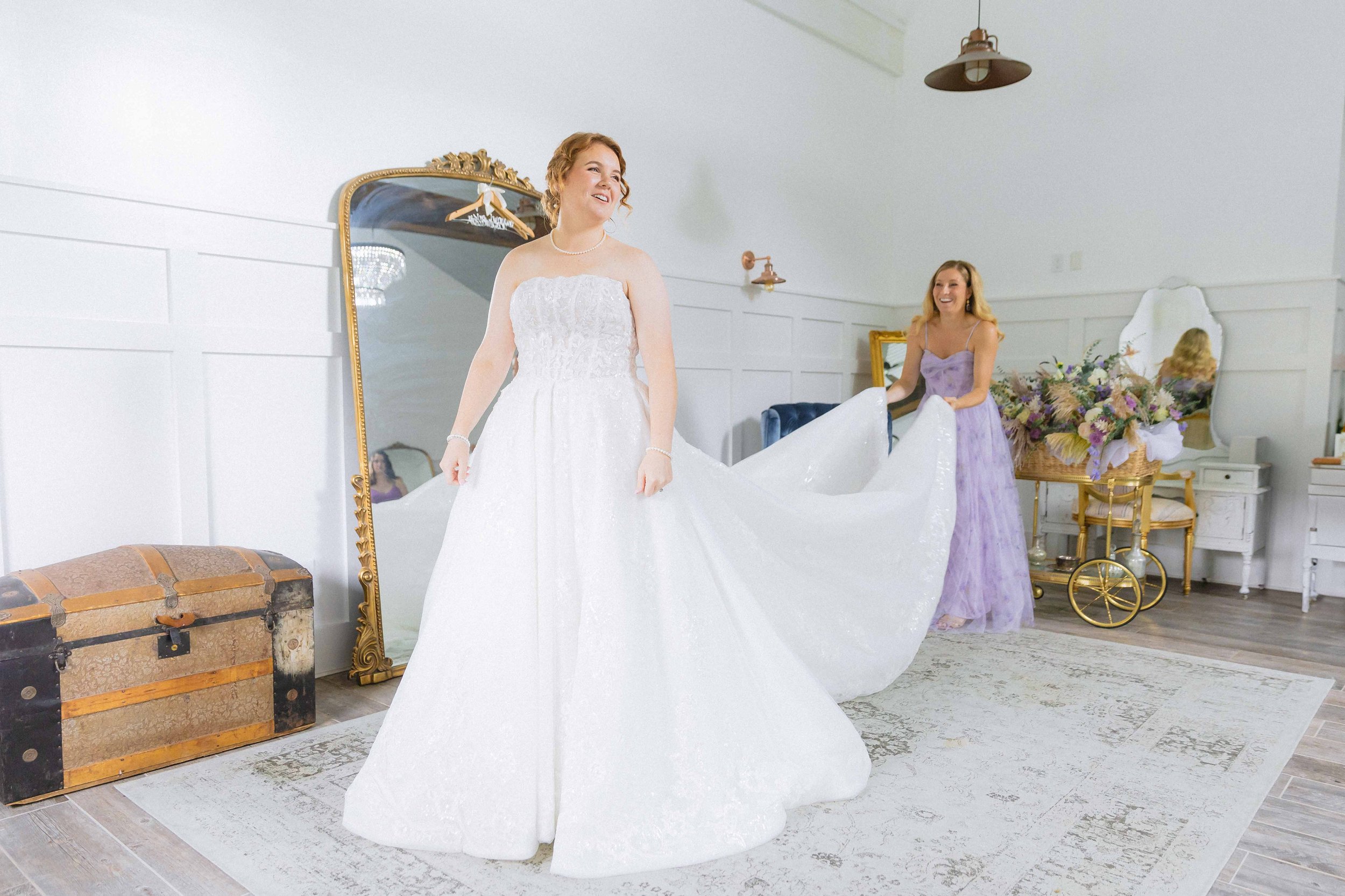 A bride in a white wedding gown being assisted by a bridesmaid in a purple dress.