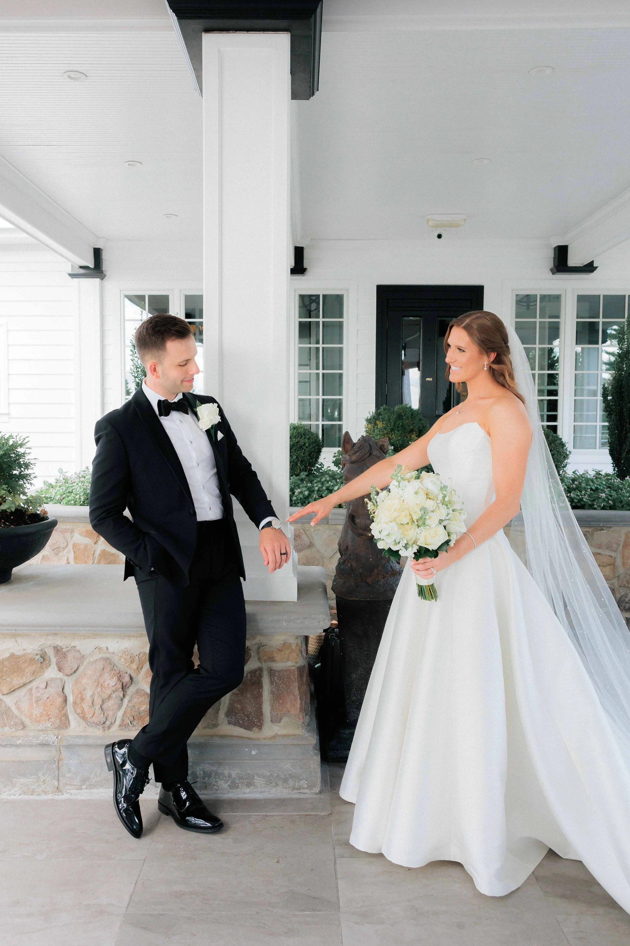Bride and groom during their wedding, standing outside a white house with black door, exchanging rings or sharing a moment.