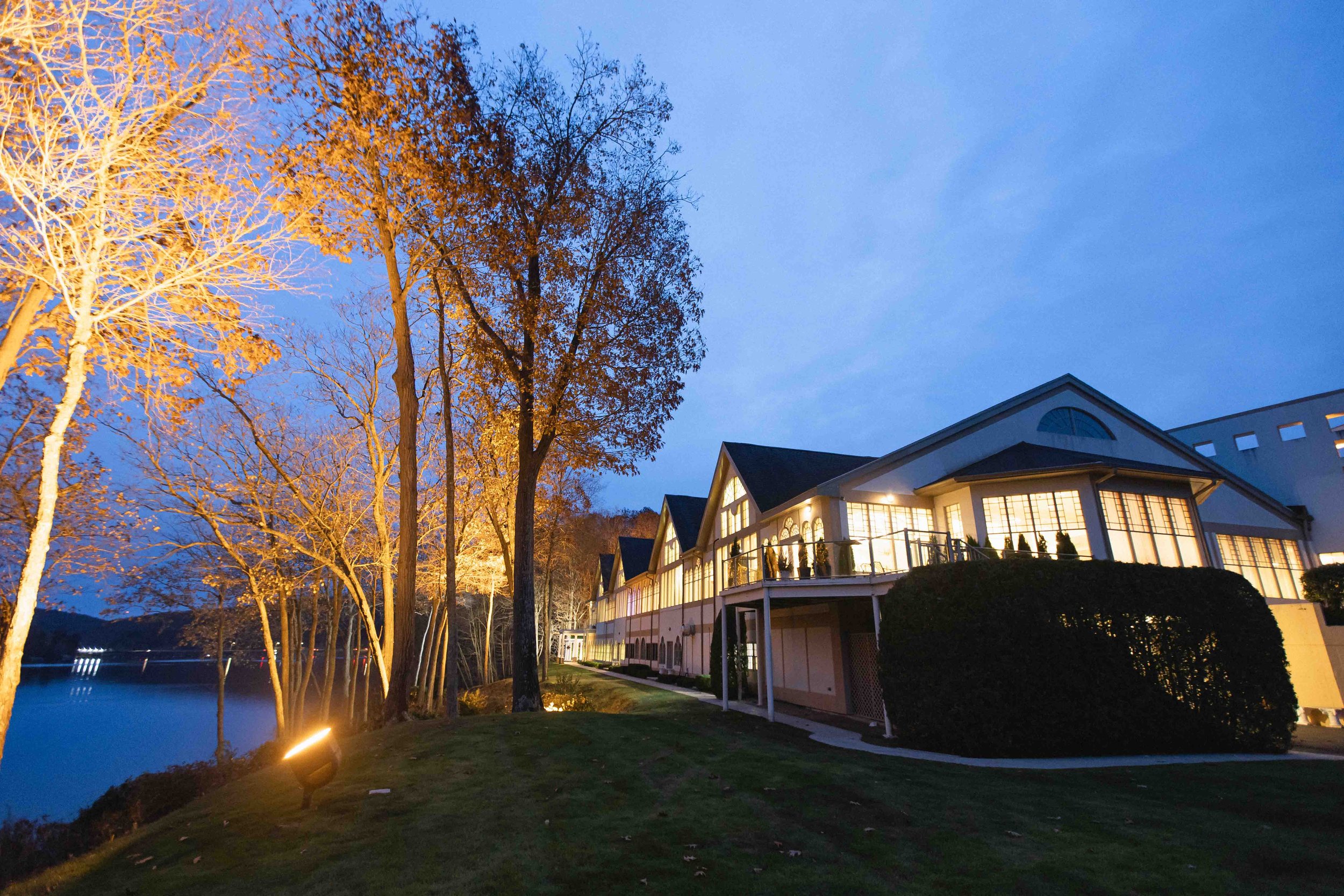 Exterior evening shot right at dusk of lakeside building overlooking lake zoar with beautifully lit trees during outdoor fall ceremony at the waterview wedding venue in monroe ct