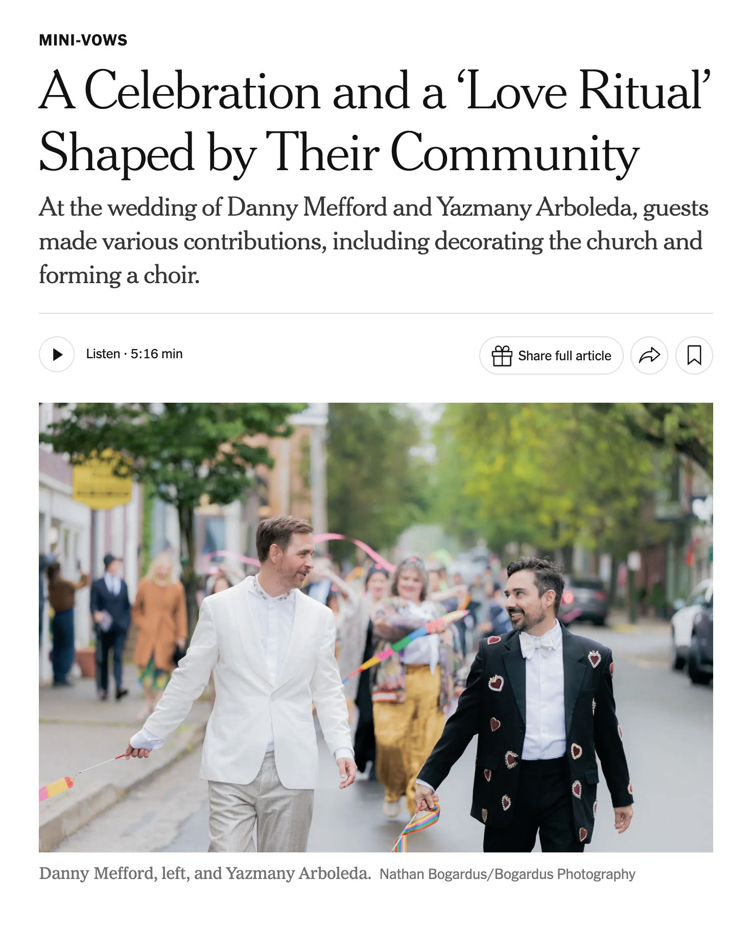 Two men, one in a white suit and one in a black suit with heart patterns, walk holding hands during a celebration or parade on a city street. They are surrounded by onlookers and participants, some holding rainbow flags, with trees and buildings in the background.