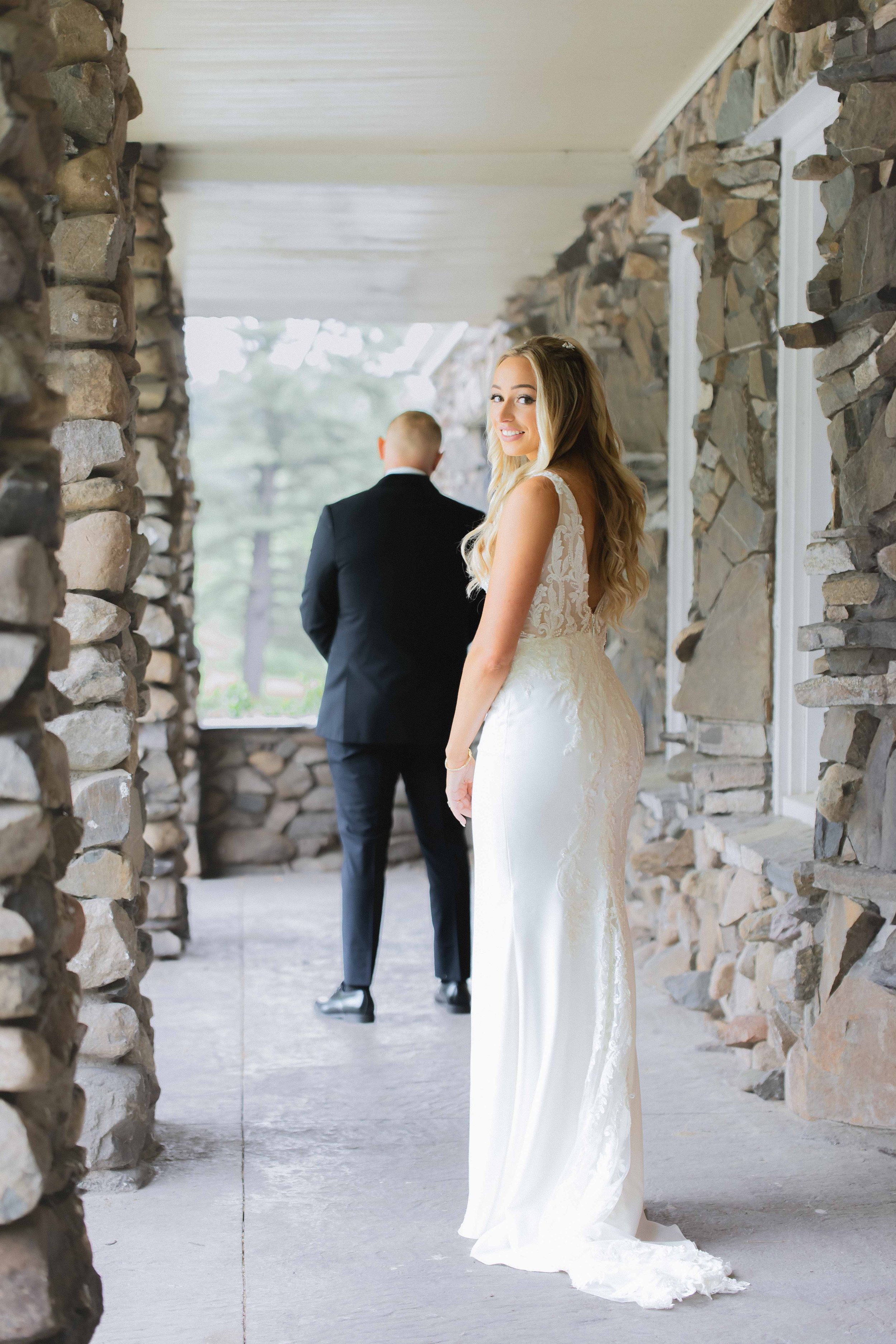 A bride in a white wedding dress smiling at the camera while standing on a stone-covered porch with a stone wall, with a man in a black suit standing facing away in the background.