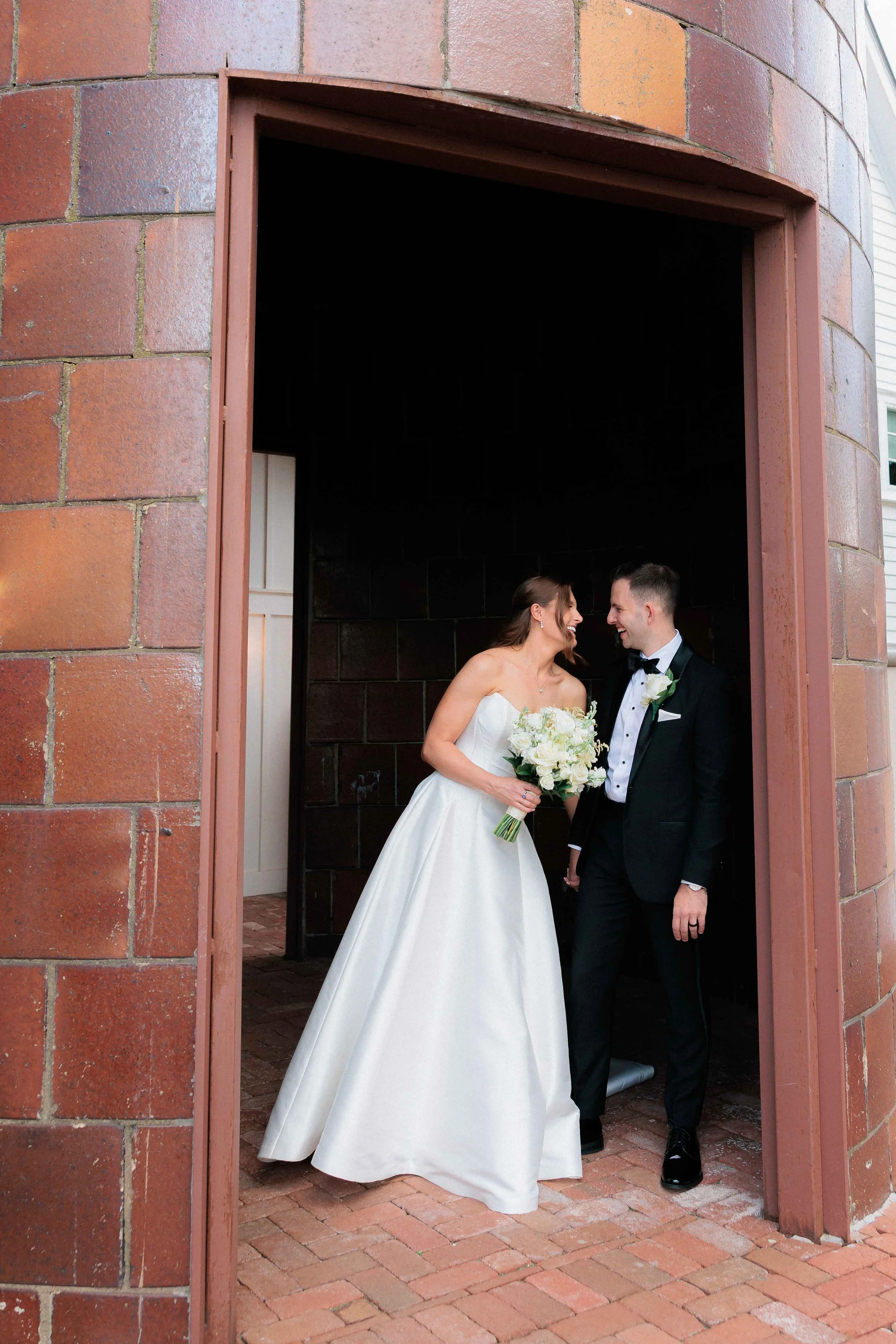 A bride and groom smiling at each other, standing in a brick archway. The bride is wearing a white strapless wedding gown and holding a bouquet of white flowers. The groom is dressed in a black tuxedo with a bow tie and a white boutonniere.
