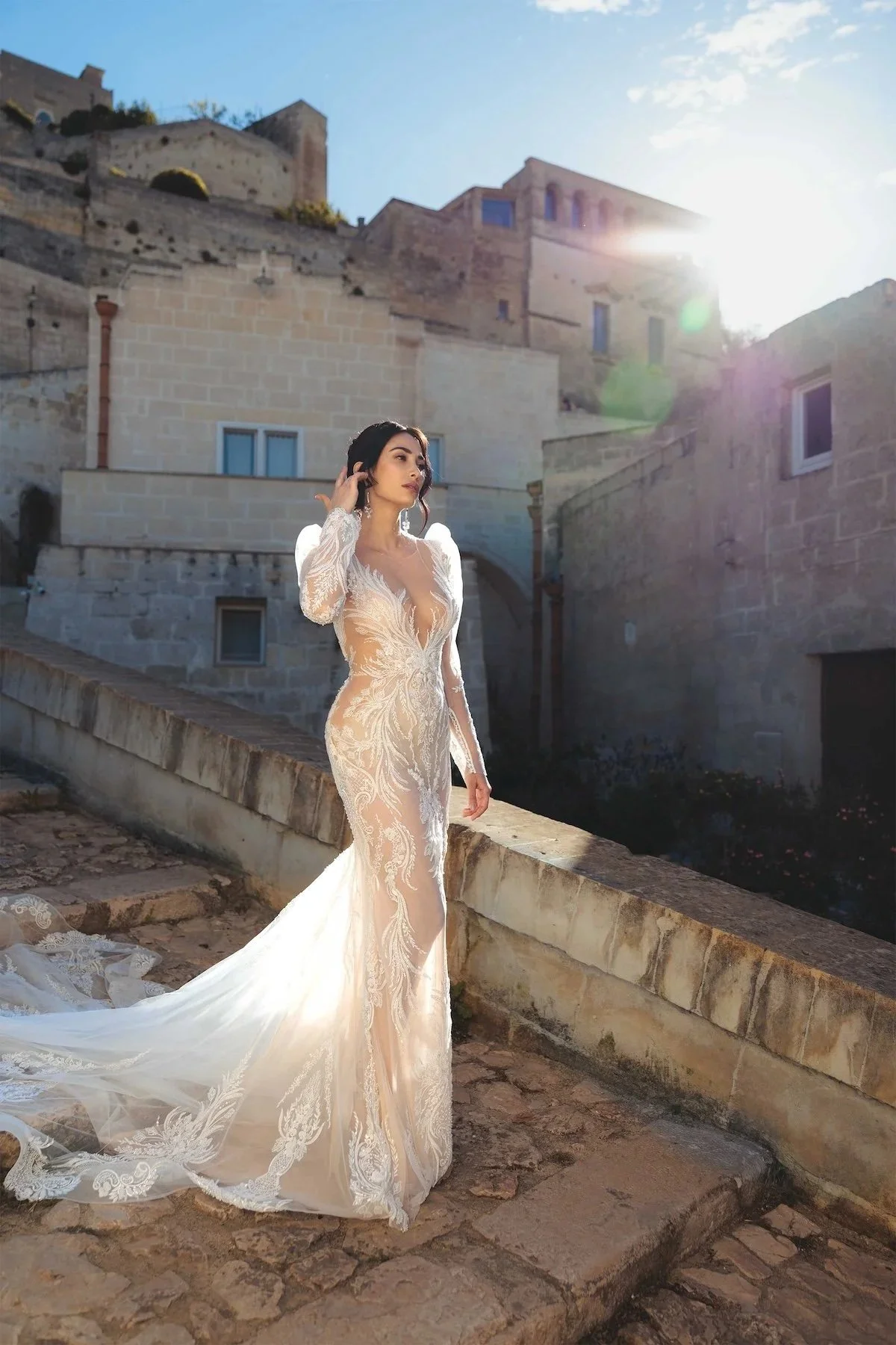 A woman in an intricate, sheer wedding dress with lace details standing outdoors on a stone walkway with old stone buildings in the background. The sun shines brightly overhead, creating a lens flare.
