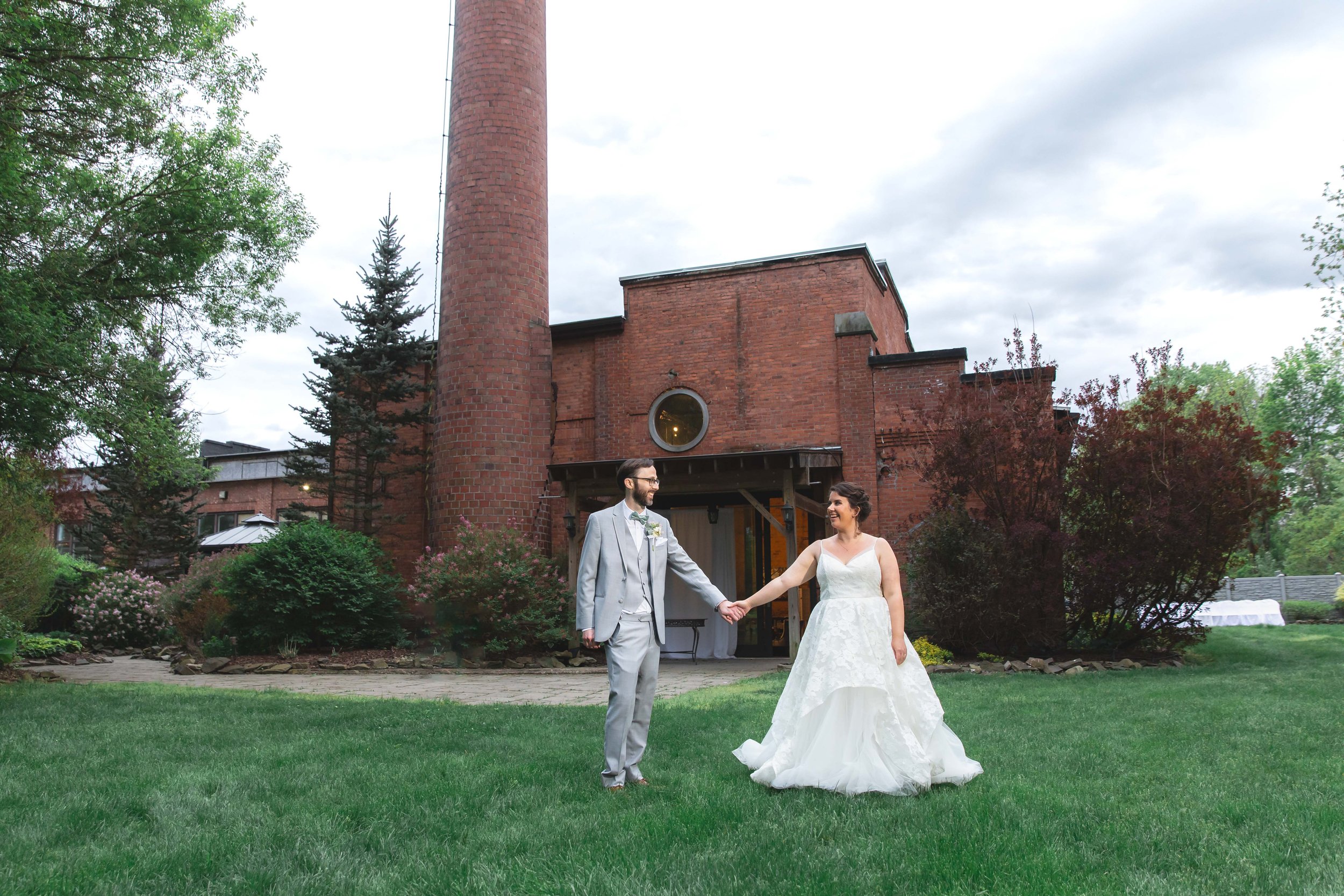 Bride and groom hold hands and pose in garden in front of industrial chic bridal suite in summer at Saugerties Performing Arts Factory wedding venue in upstate NY