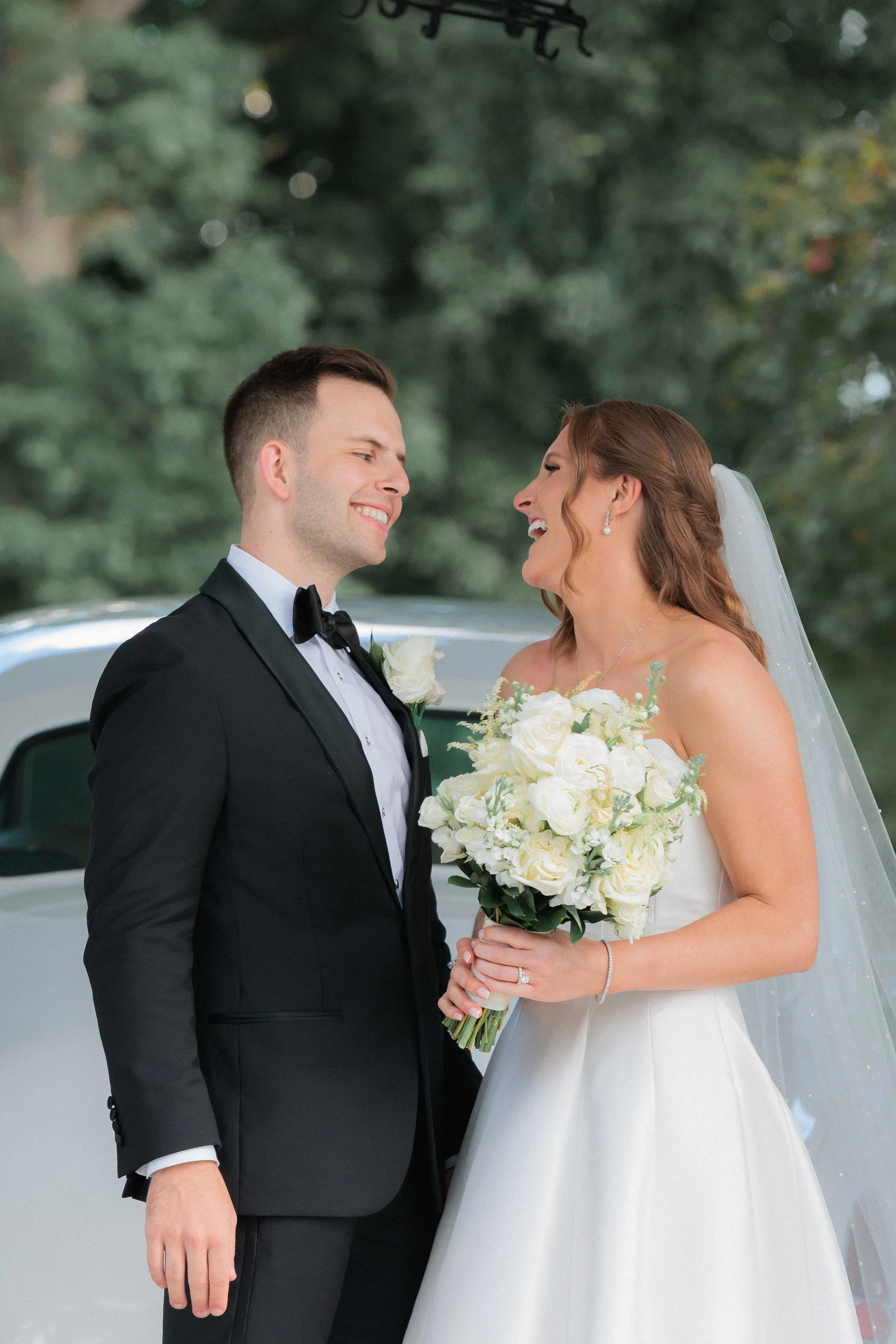 A bride and groom on their wedding day, smiling at each other outdoors. The bride is holding a bouquet of white flowers and wearing a white wedding gown with a veil. The groom is dressed in a black tuxedo with a bow tie, standing close to the bride.