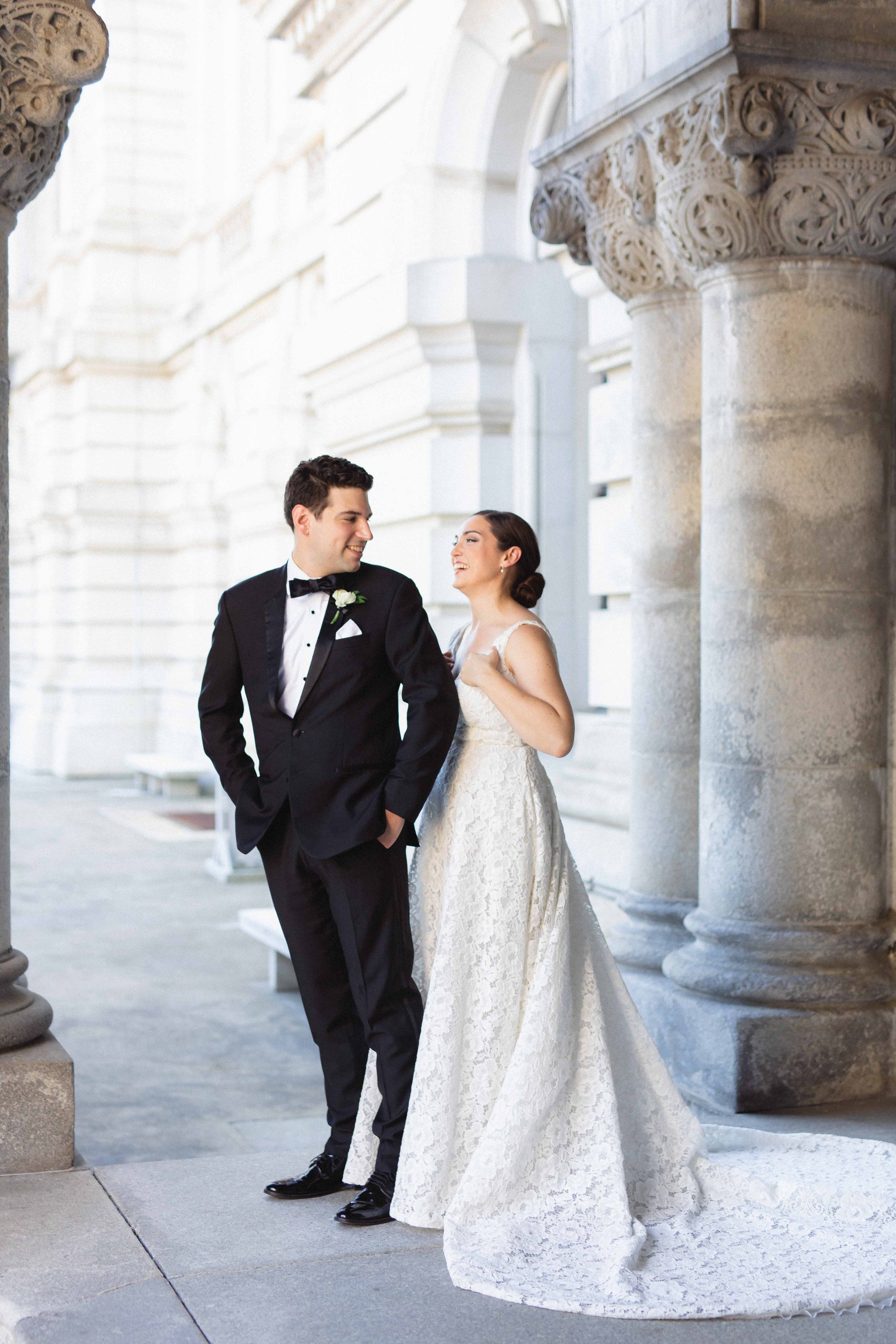 A bride and groom smiling at each other outside a historic building with stone columns