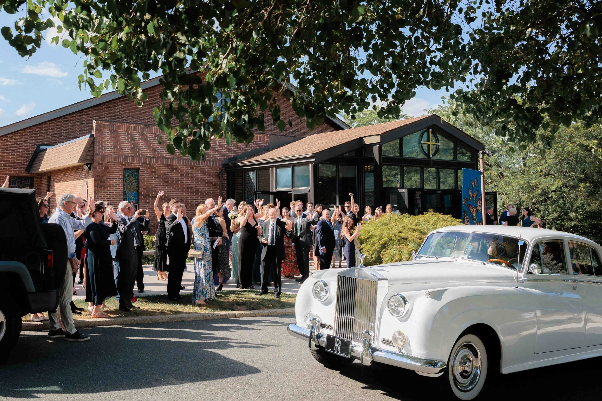 A vintage white Rolls Royce parked outside a modern brick church, with a group of well-dressed people waving and celebrating in the background on a sunny day.