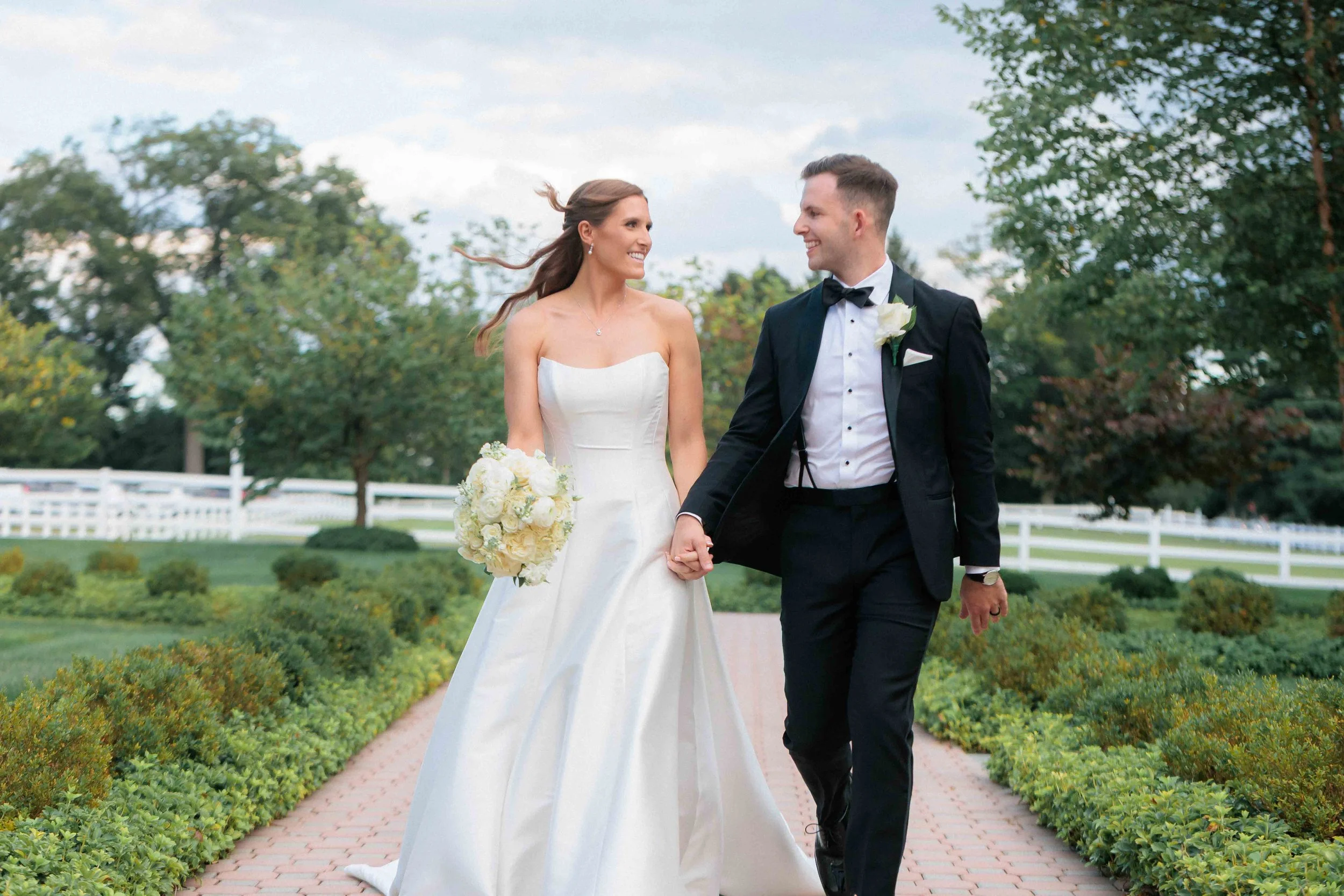 A newlywed couple walking hand-in-hand outdoors, smiling at each other, surrounded by greenery and trees.