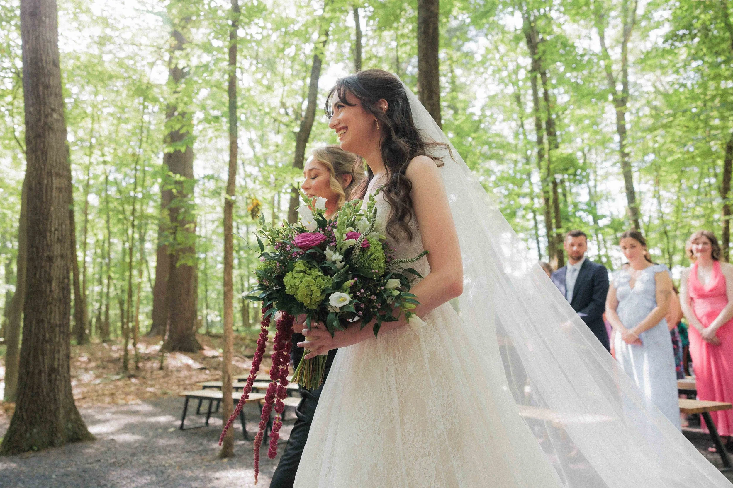 A bride holding a bouquet of flowers during an outdoor wedding ceremony in a forest, with her eyes closed and smiling, while guests stand in the background.