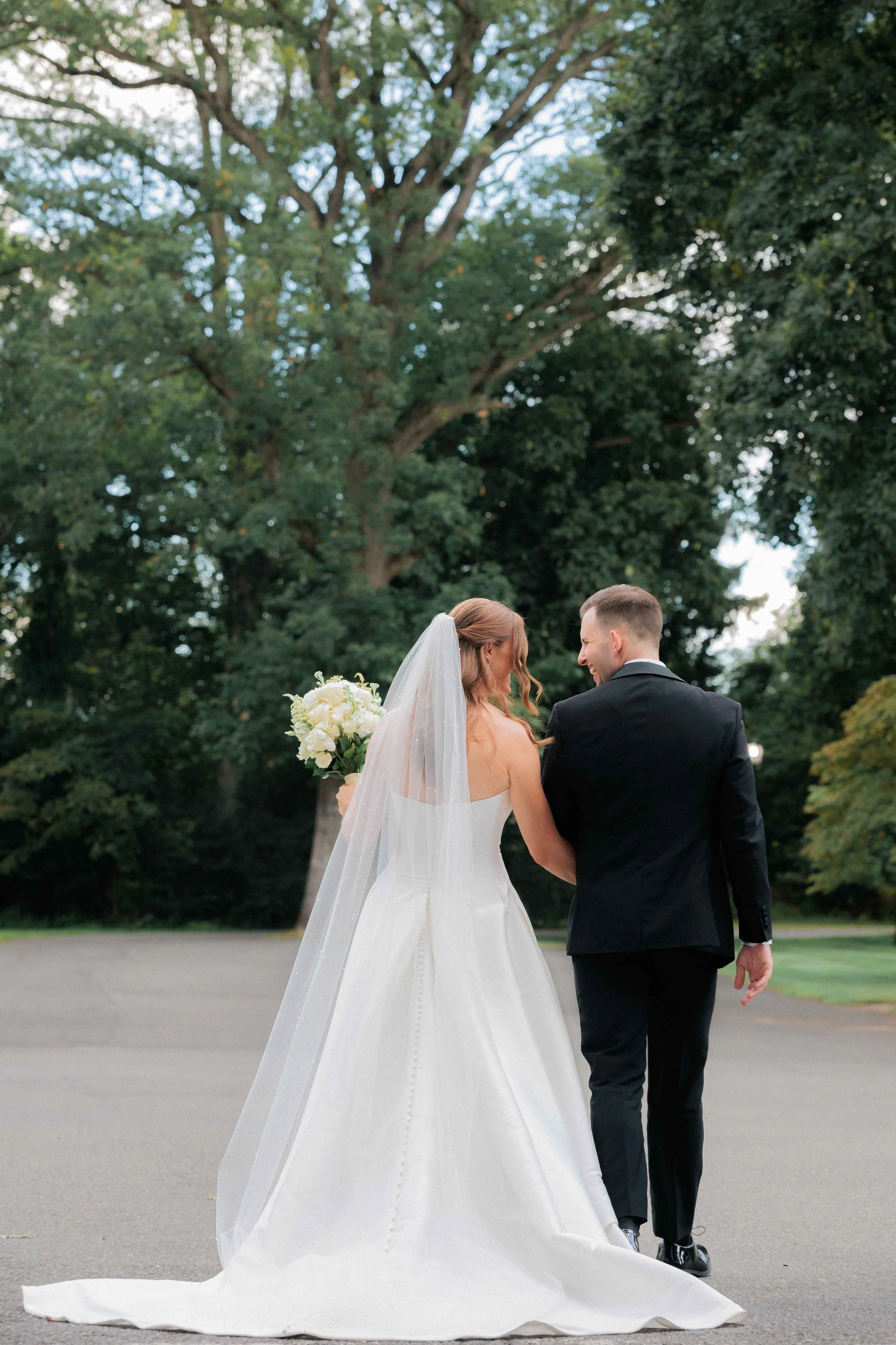 Bride and groom walking together outdoors, bride holding a bouquet of white flowers, surrounded by green trees.
