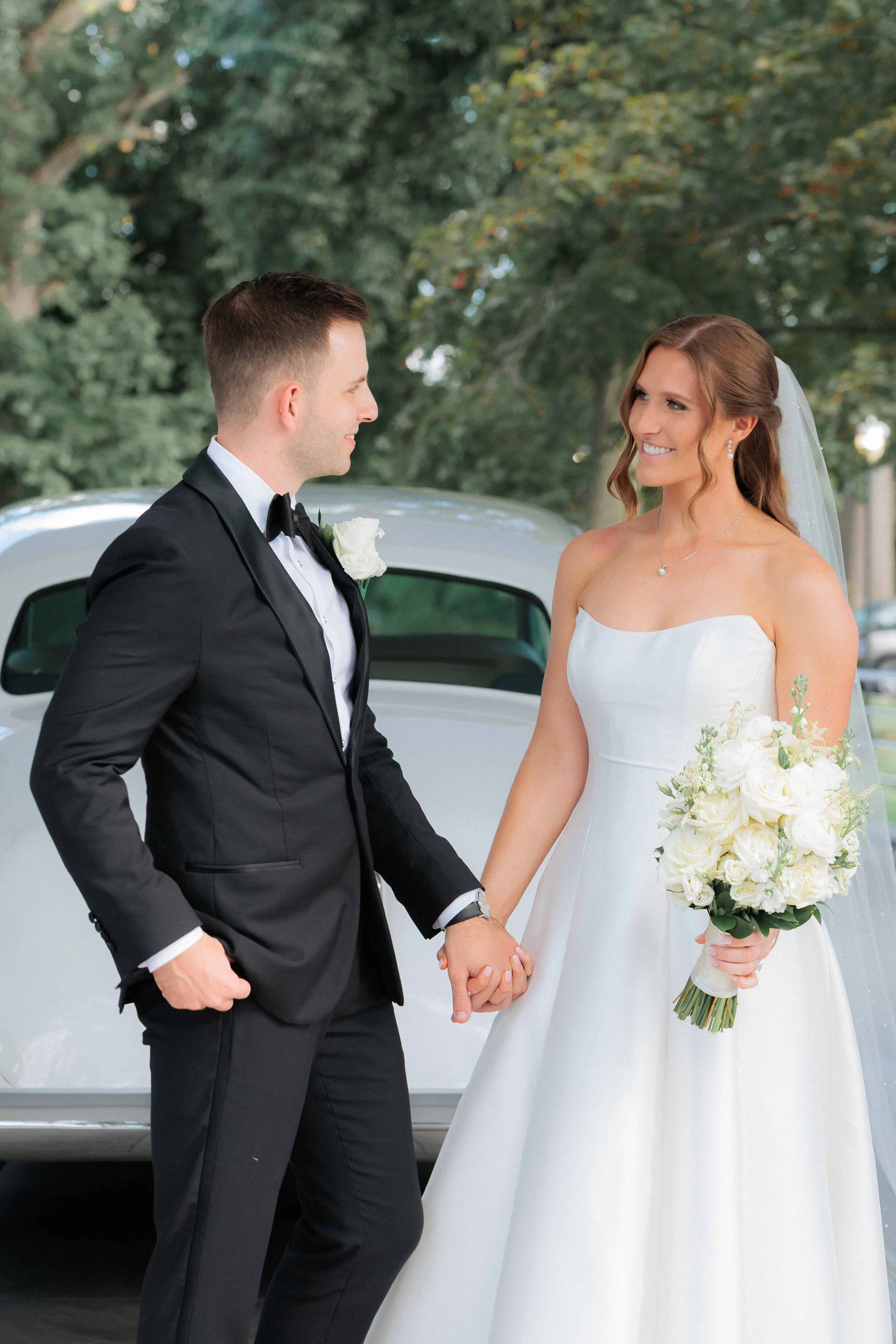 A bride and groom holding hands and smiling at each other outdoors next to a vintage white car, with trees in the background.