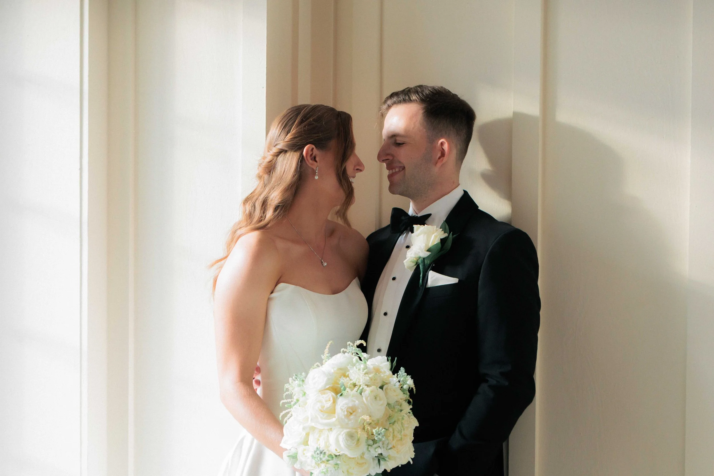 A bride and groom standing close together, smiling, with the bride holding a bouquet of white roses, in a softly lit indoor setting.